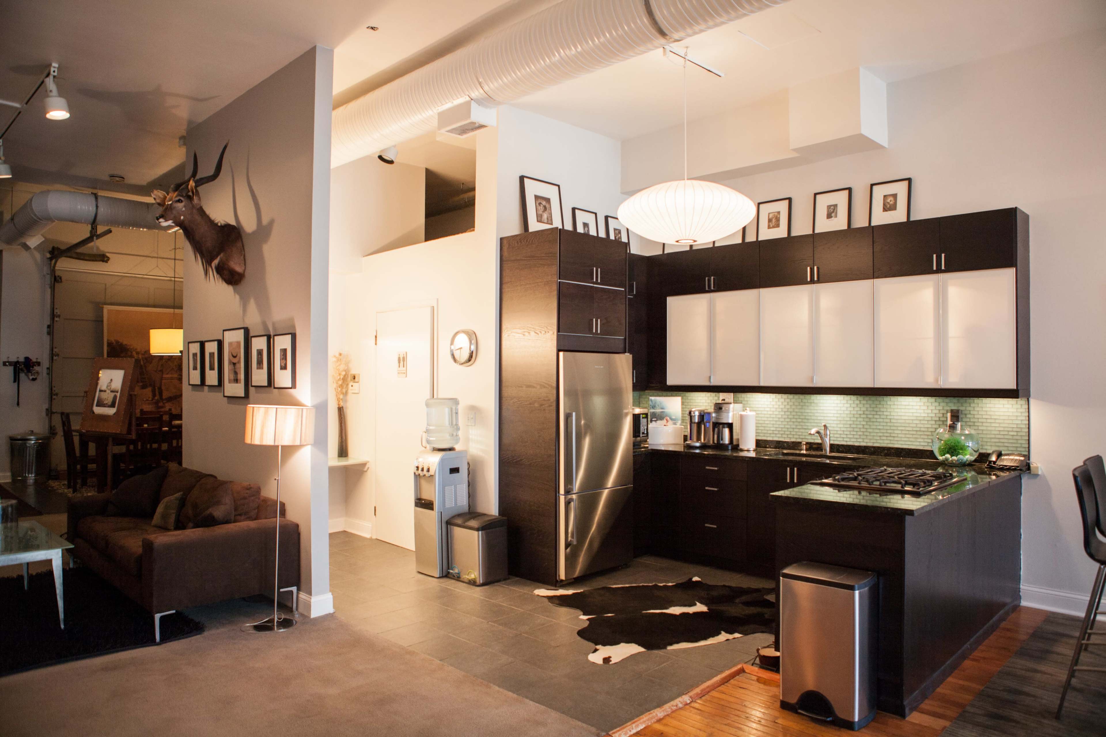 The image shows a modern open-concept apartment featuring a kitchen with dark cabinetry, a living area with a brown sofa, and decorative wall-mounted frames.