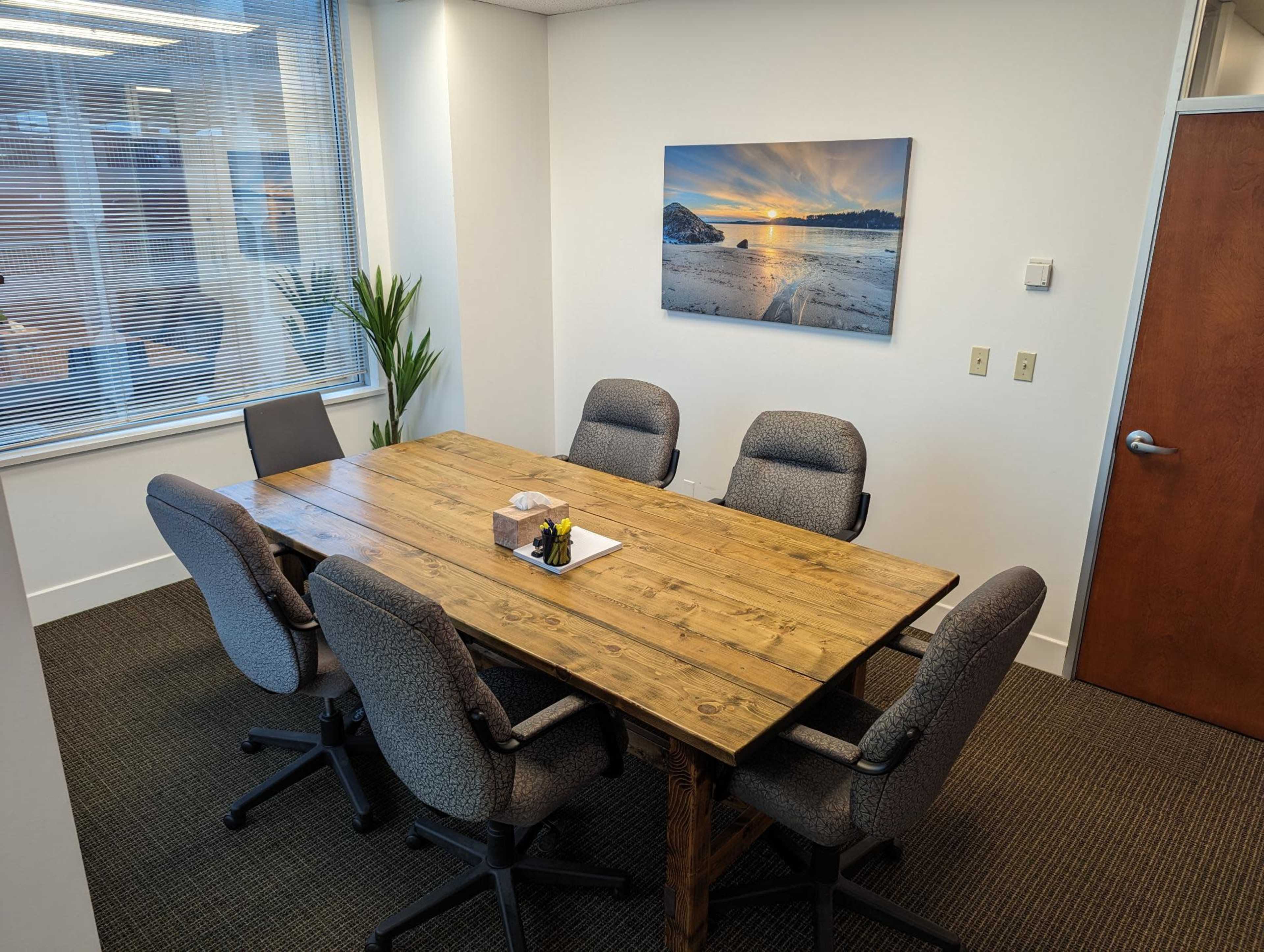 A conference room features a large wooden table surrounded by six gray office chairs, with large windows and a landscape photo on the wall.