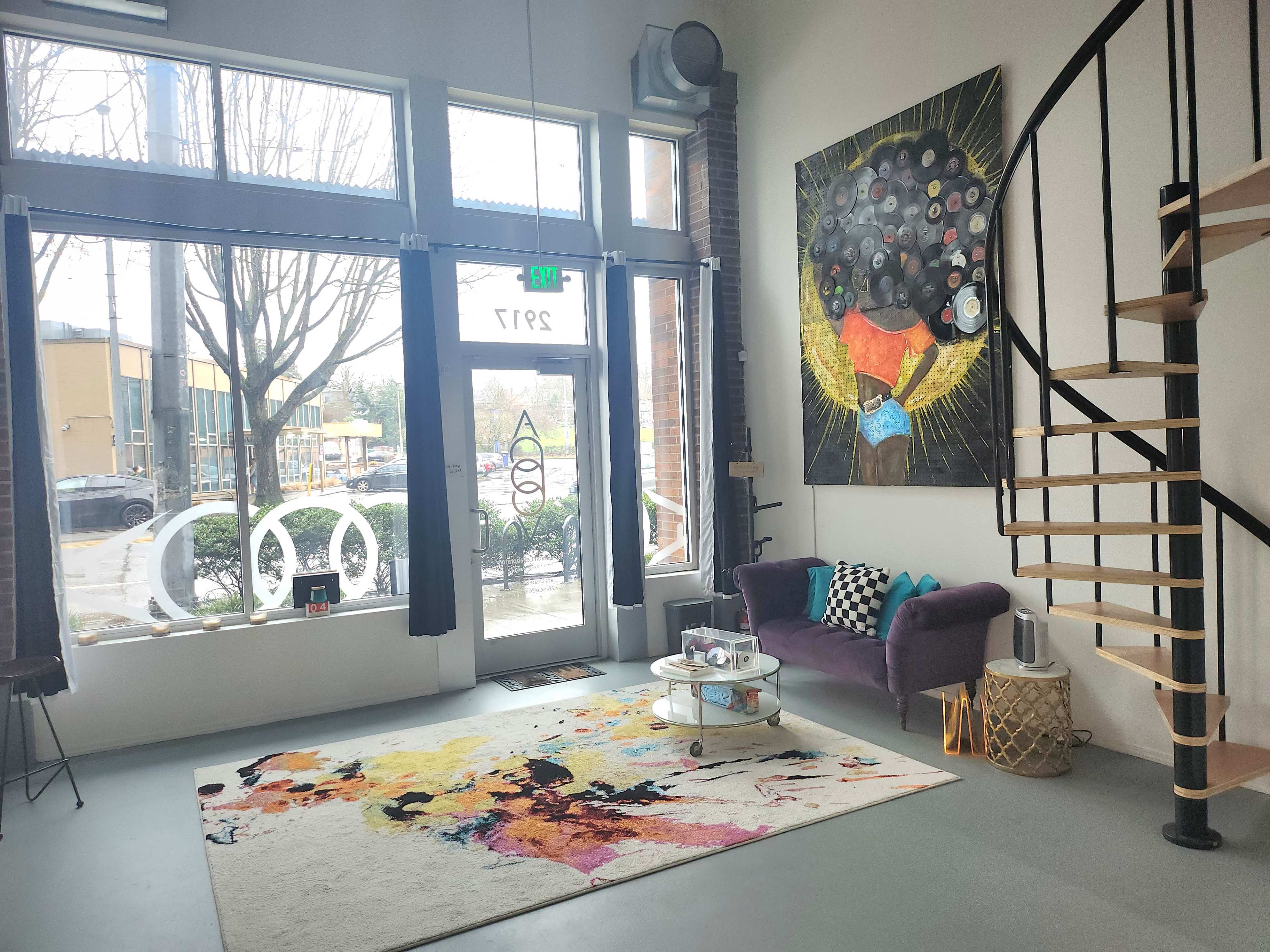 A spacious, modern room with large windows, a purple sofa, a colorful area rug, and a staircase leading to an upper level, accompanied by a vibrant wall art piece.