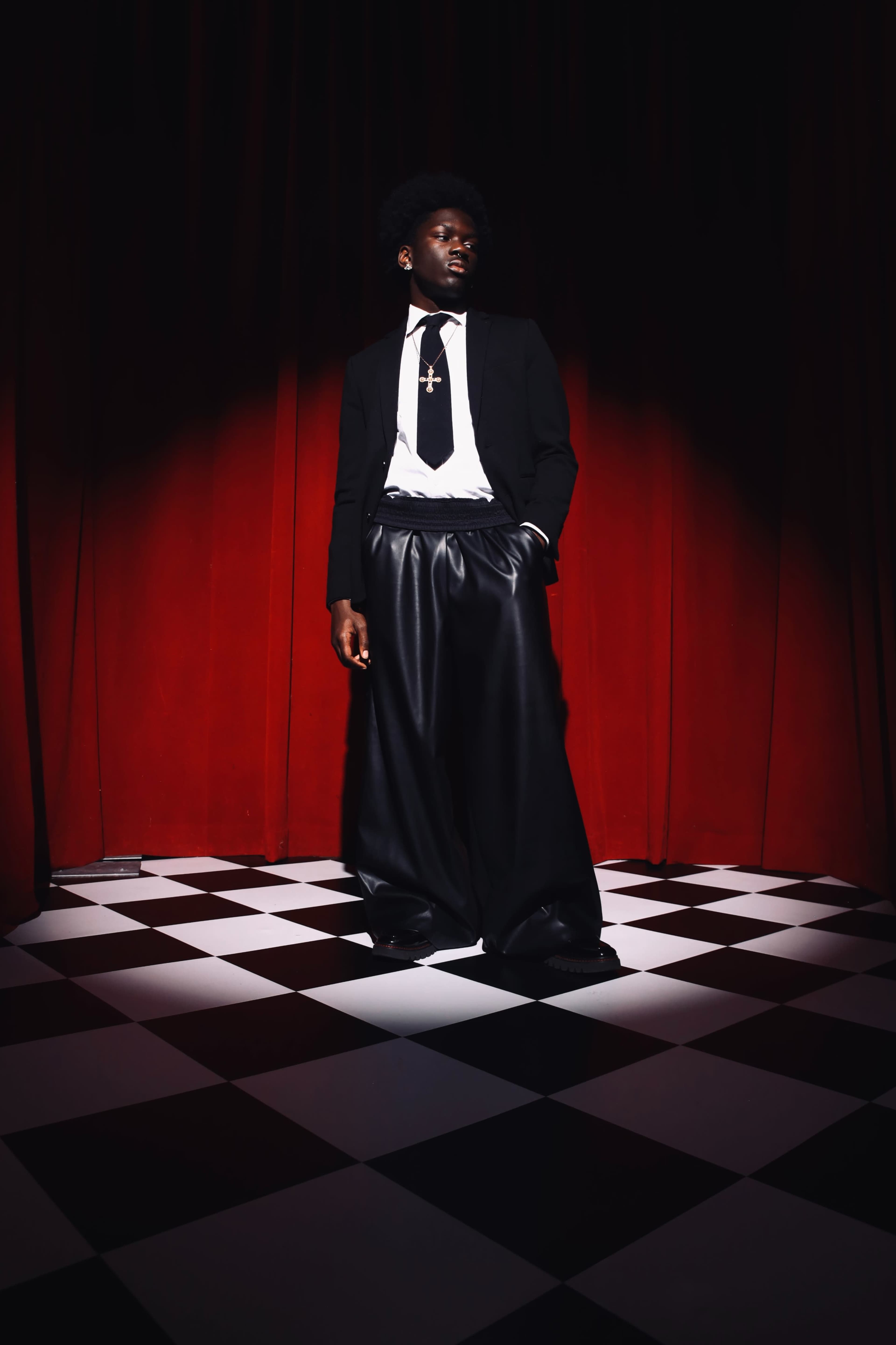 A person wearing a black suit and oversized trousers stands on a checkered floor under dramatic lighting in front of a red curtain.