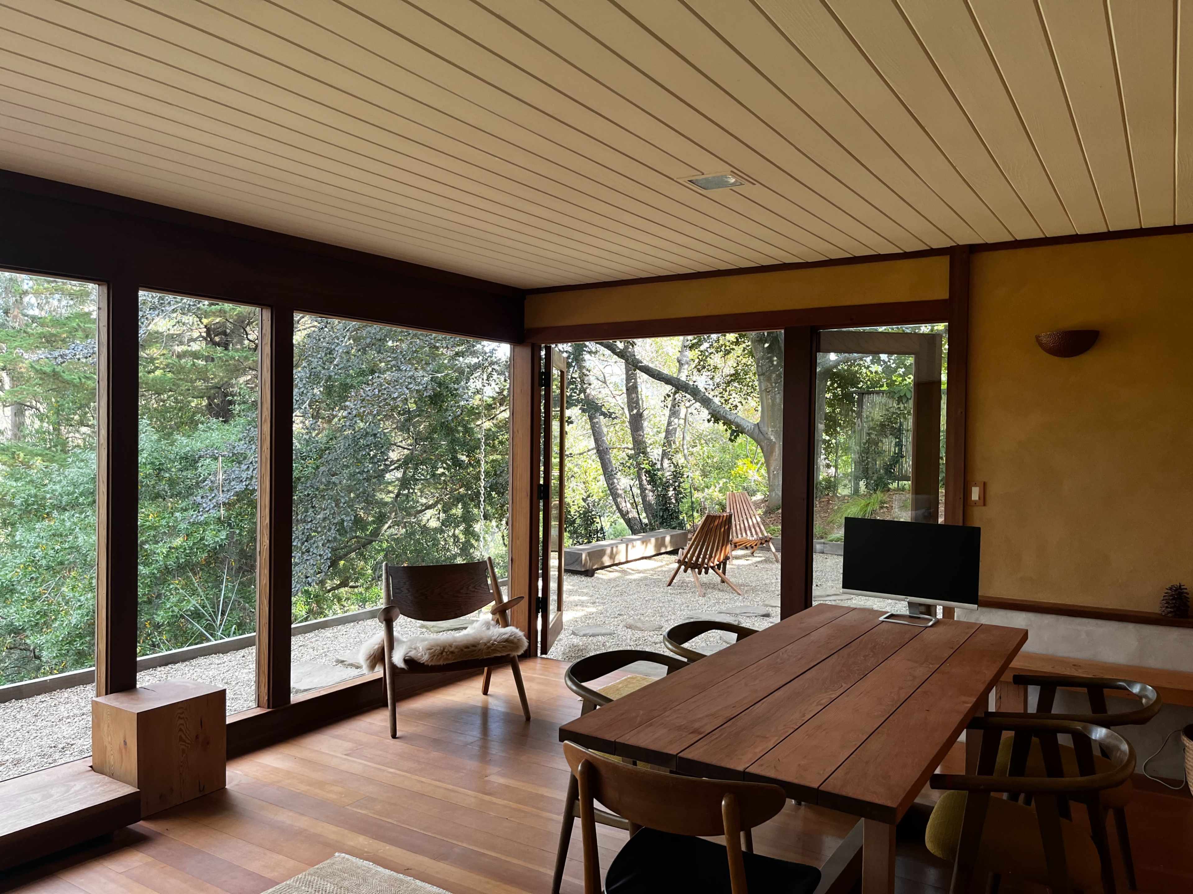 A modern, open-concept room features a wooden dining table, chairs, and large windows overlooking a natural outdoor space with trees.