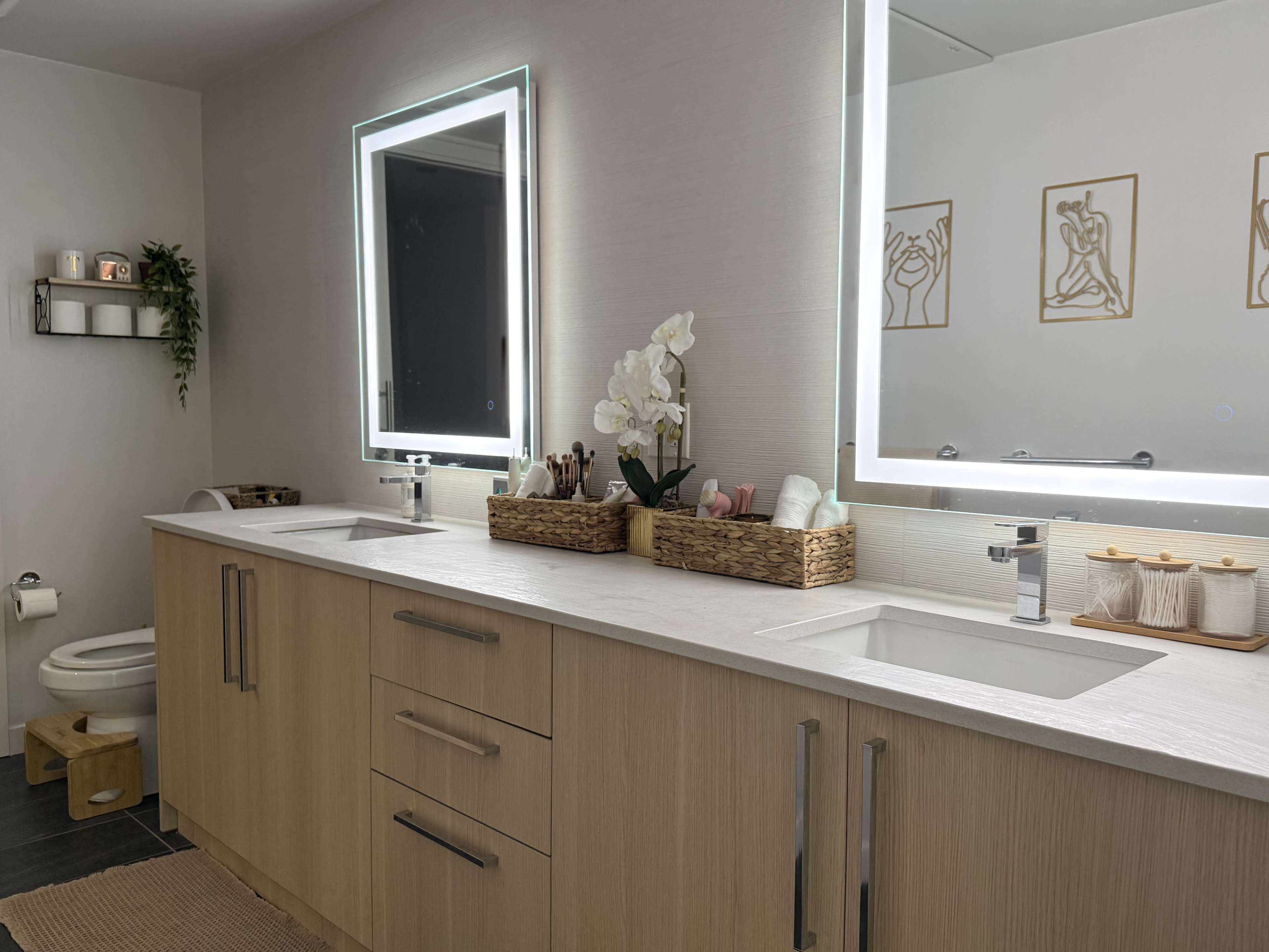 The image shows a modern bathroom with a double sink vanity, two illuminated mirrors, and organized storage baskets on the countertop.