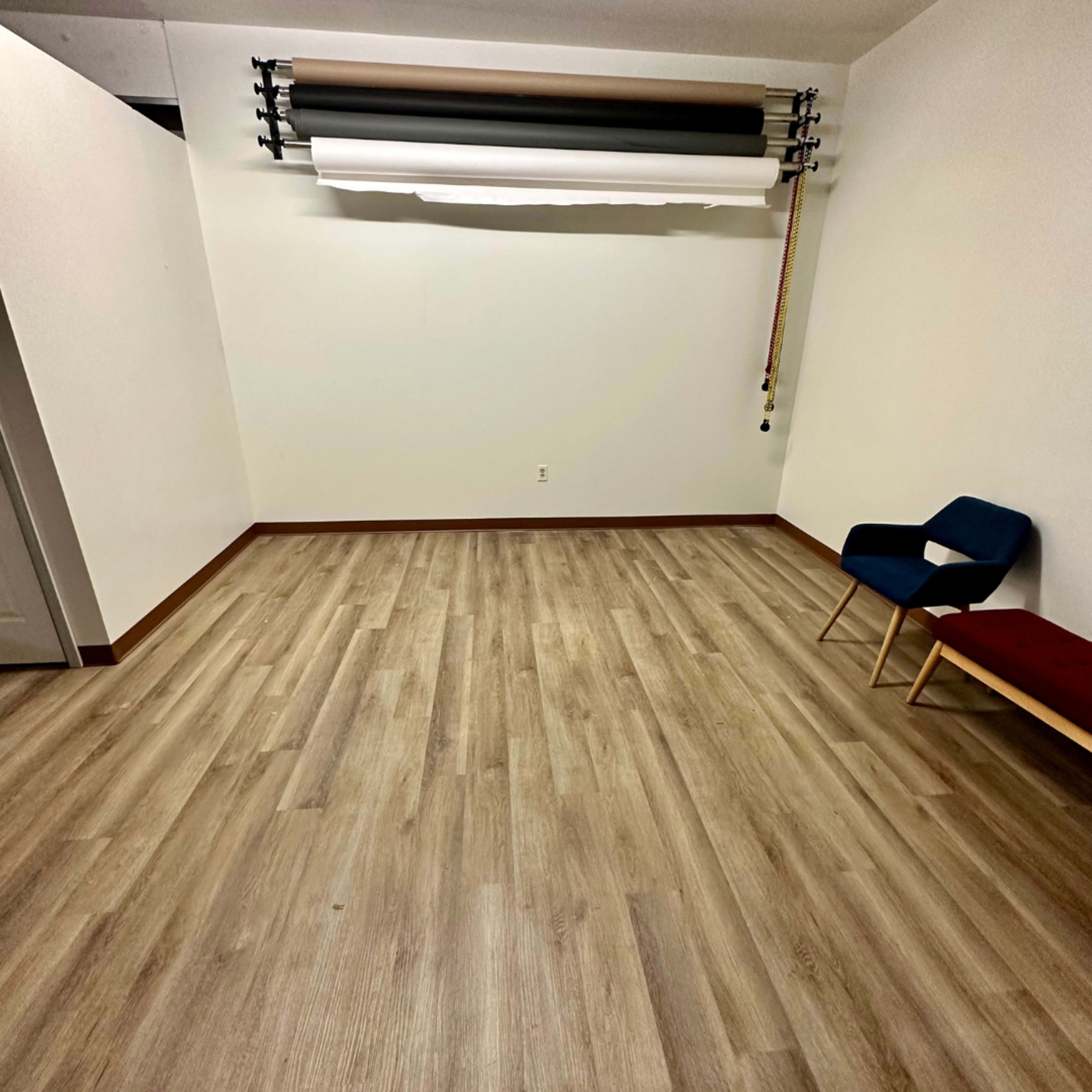 A minimalist interior room with wooden flooring, a blank white wall, and two chairs, one blue and one red, near the corner.