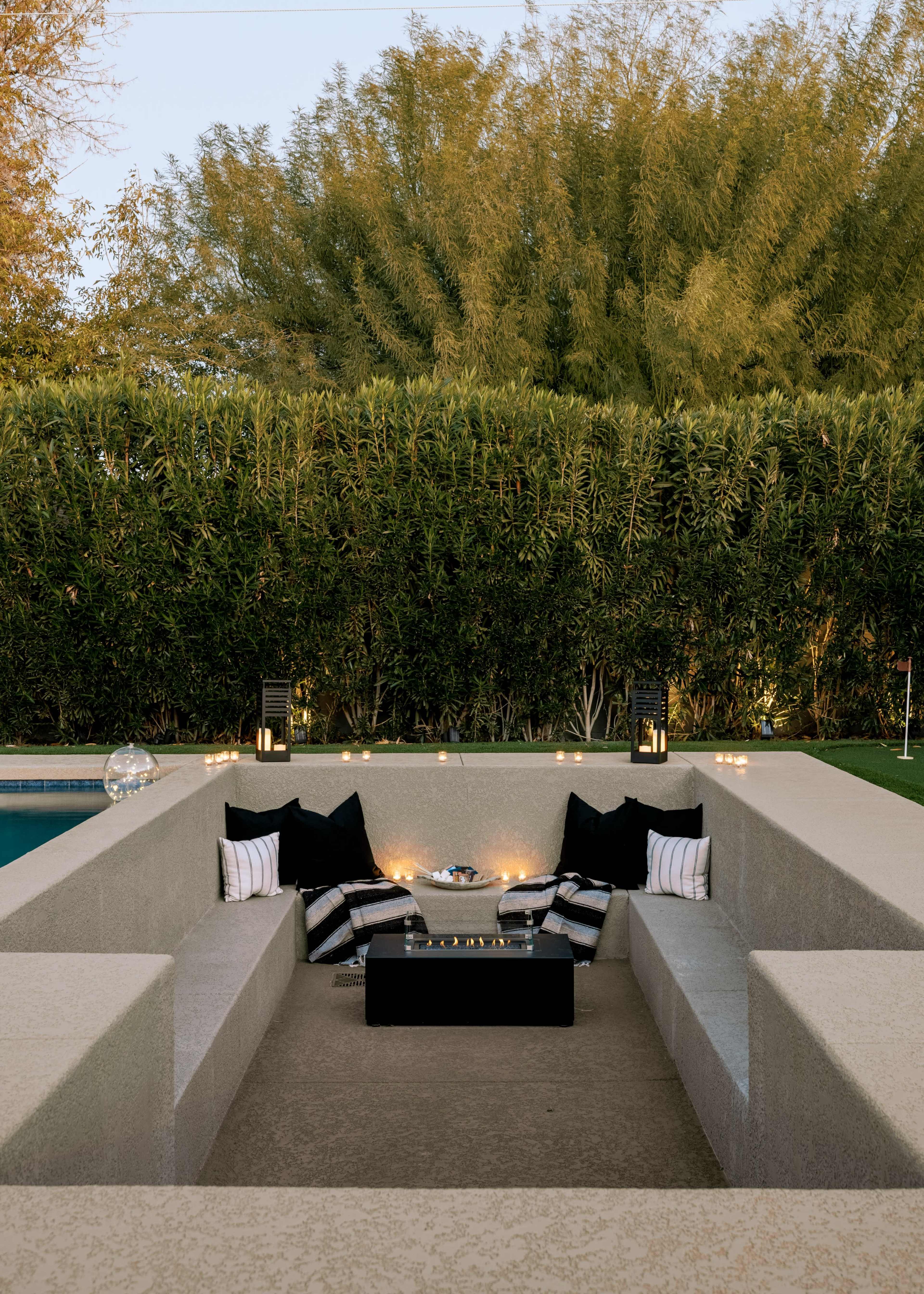 The scene features a sunken outdoor seating area with a rectangular table, surrounded by plush cushions and decorative pillows, flanked by greenery and ambient lighting.