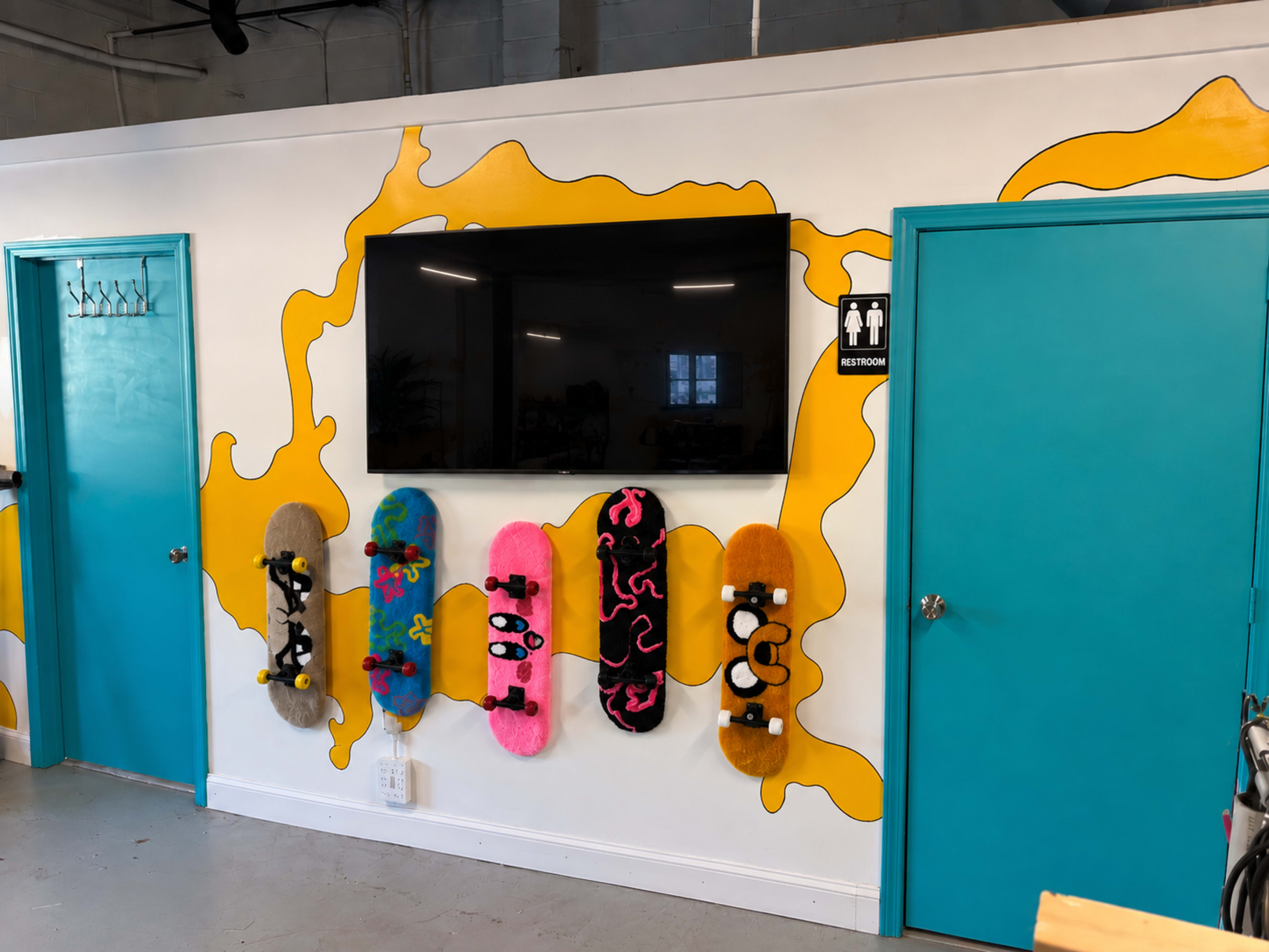 The image shows a colorful wall with a large flat-screen TV mounted above a display of six creatively decorated skateboards, flanked by two teal doors labeled with restroom signs.