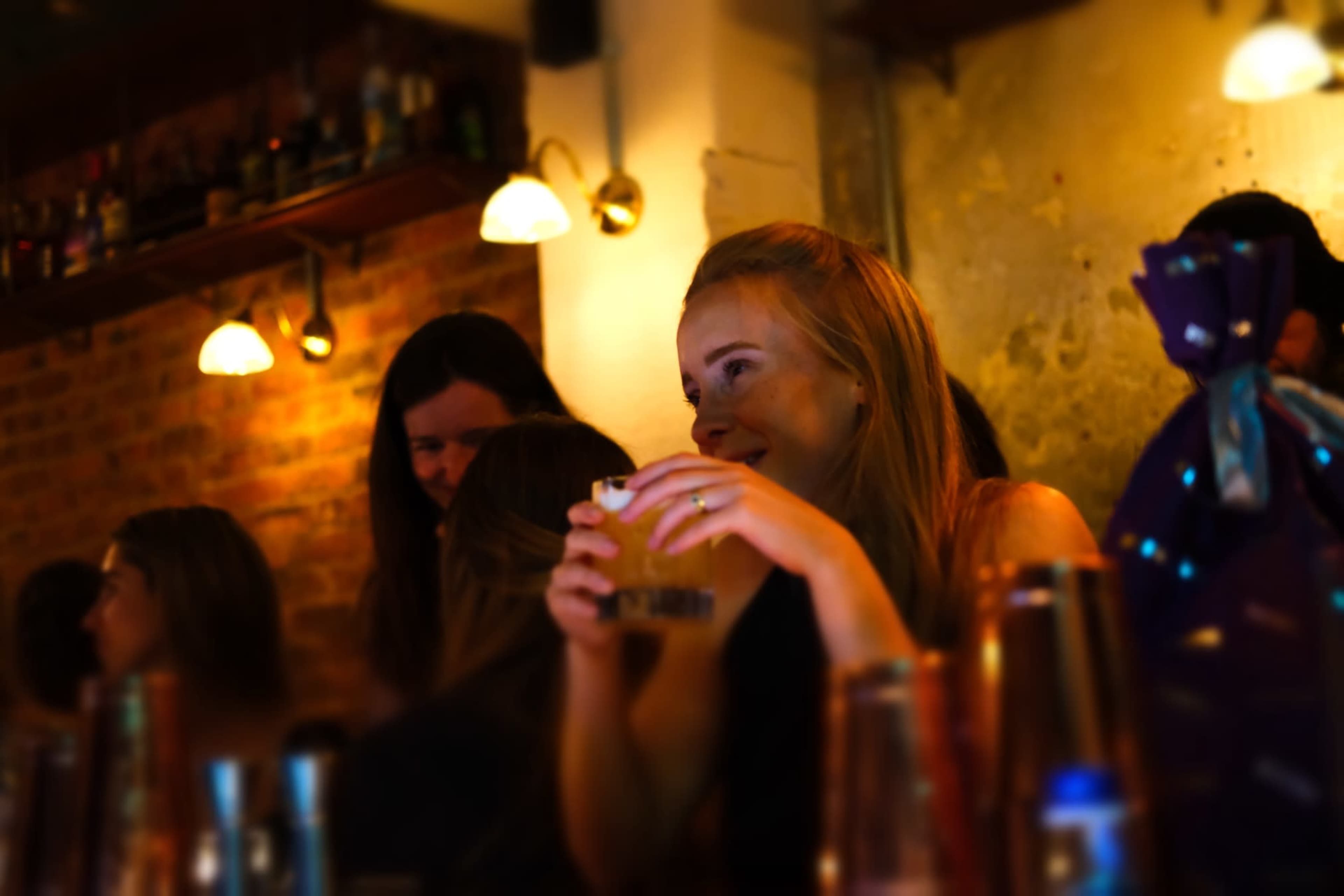 A young woman holds a drink at a dimly lit bar while people chat in the background.