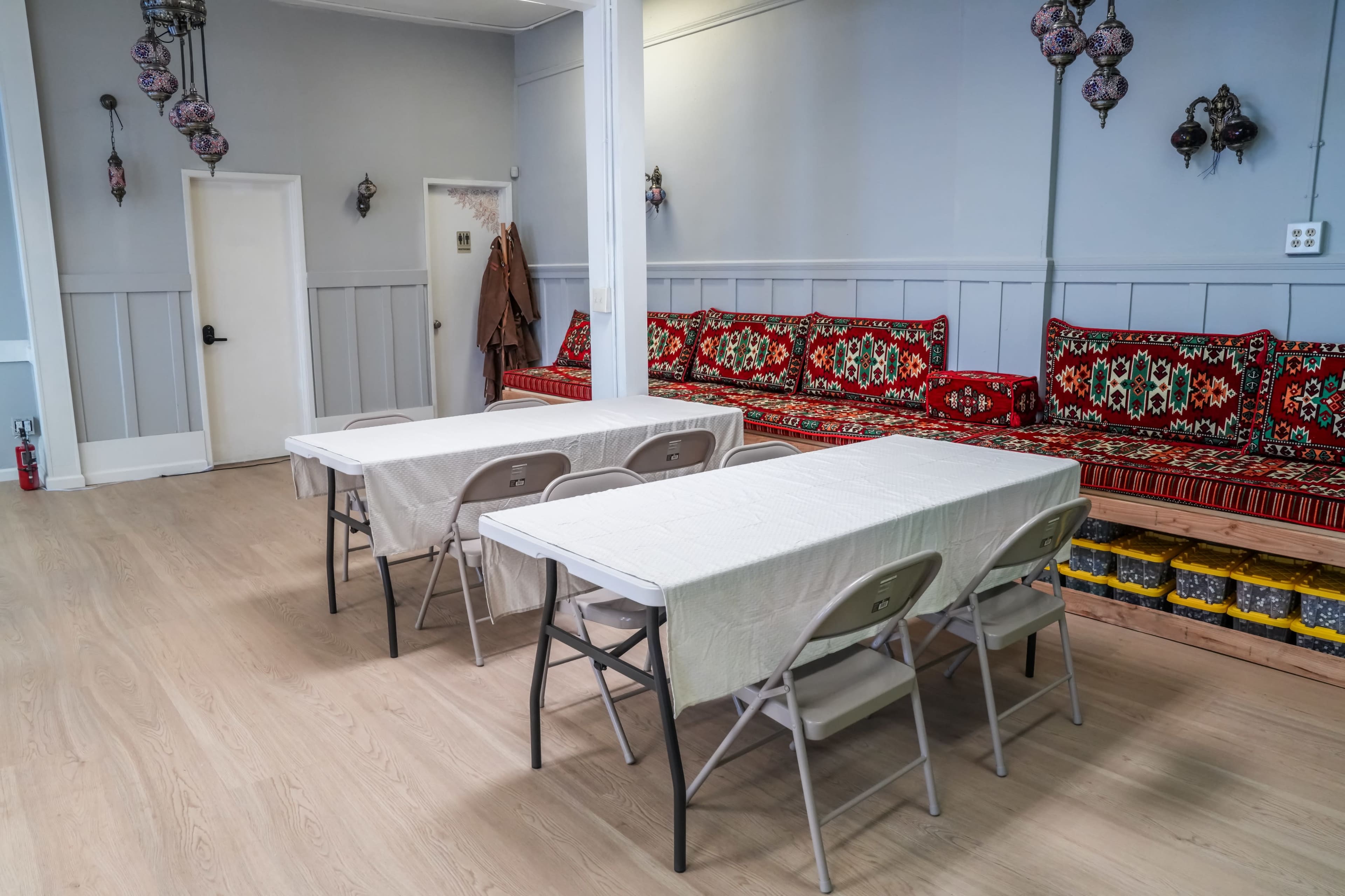 The image shows a spacious room with two long tables surrounded by folding chairs, and decorative cushions lining the walls.