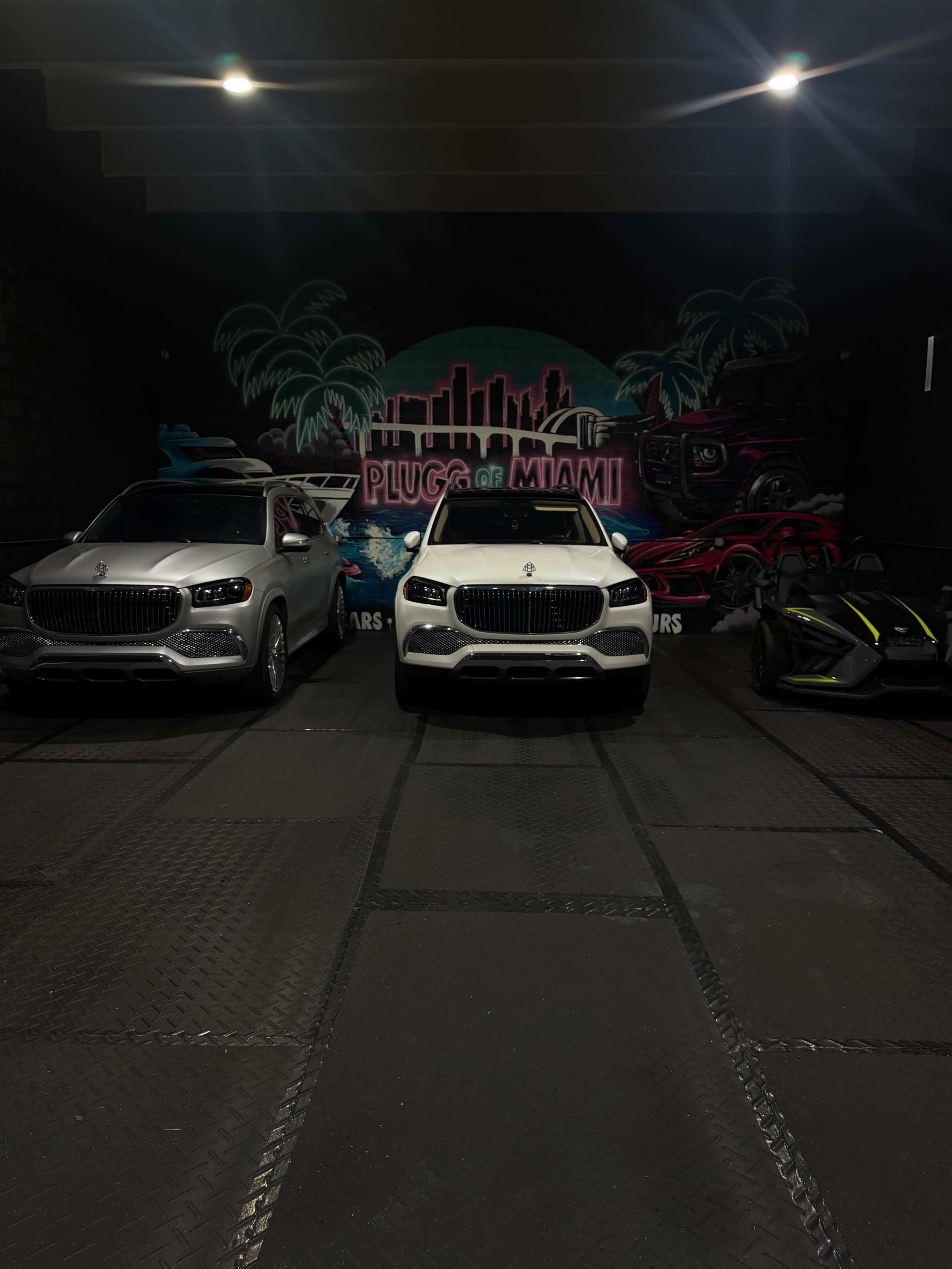 Three luxury vehicles, including two white SUVs and a black sports car, are parked in front of a colorful mural that reads "Plugg of Miami."