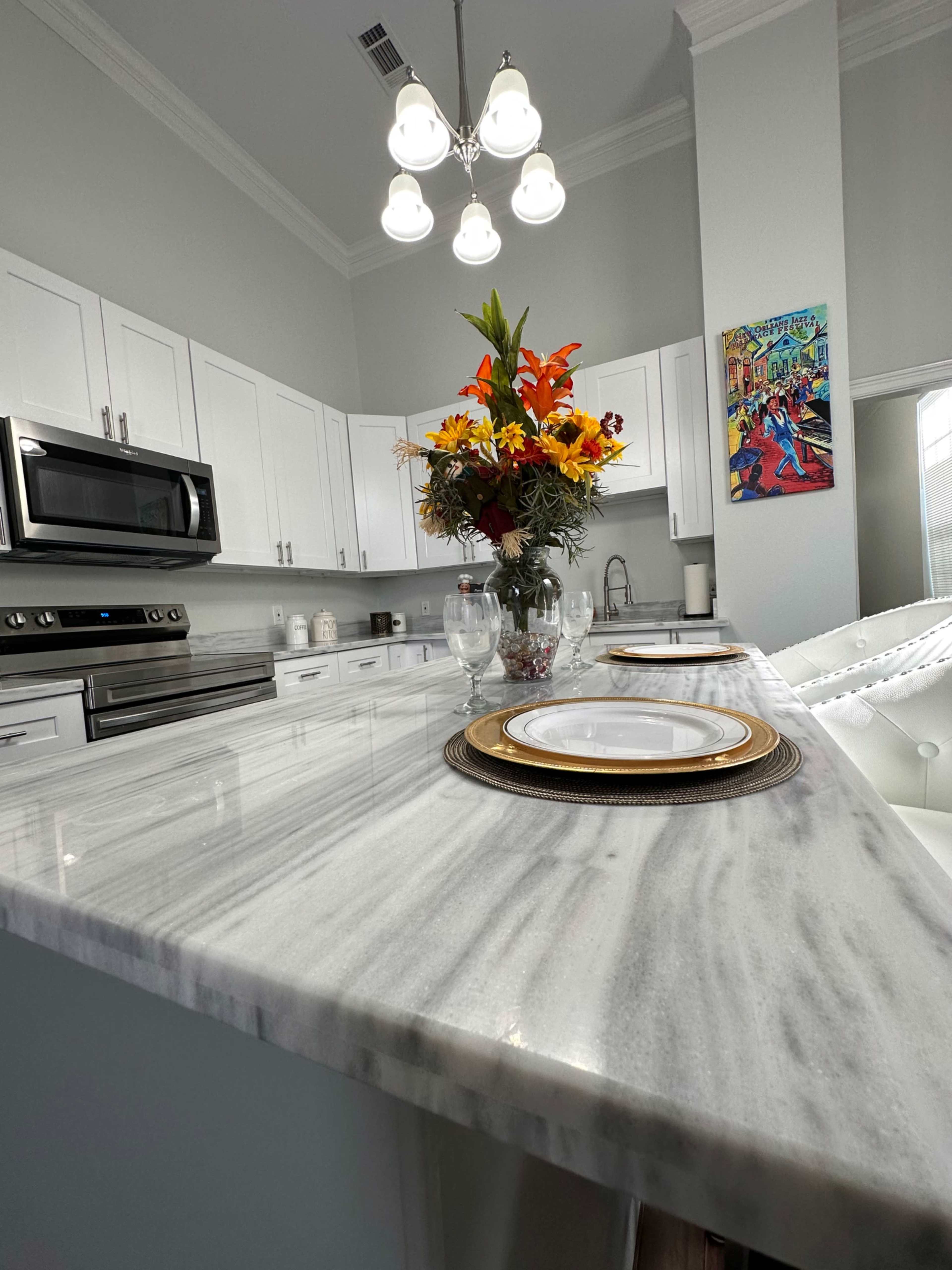 A modern kitchen features a marble island with place settings and a vase of colorful flowers, complemented by white cabinetry and stainless steel appliances.