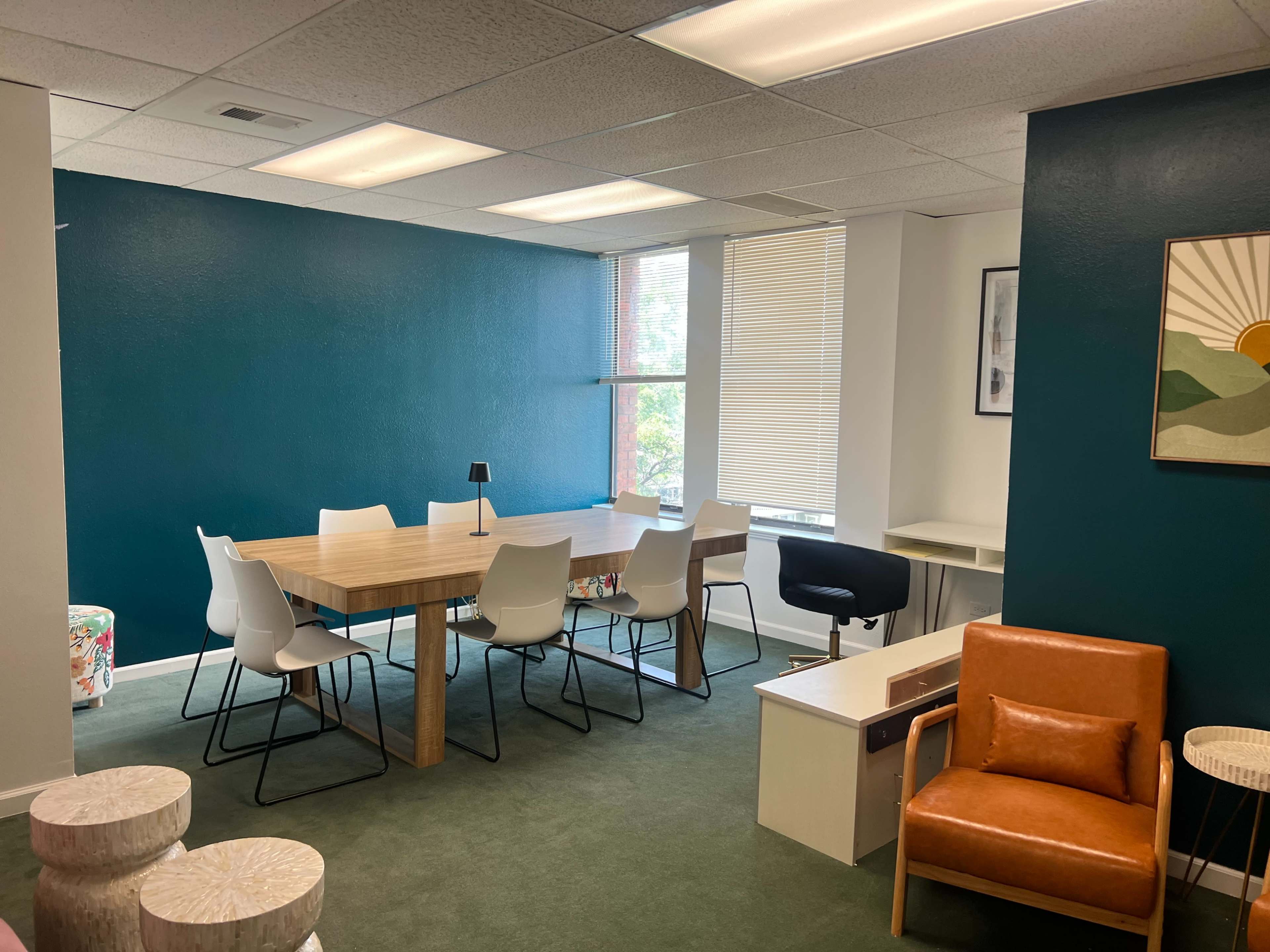 The image shows a modern office space featuring a large wooden table surrounded by white chairs, a brown armchair, and a small desk against a teal wall.