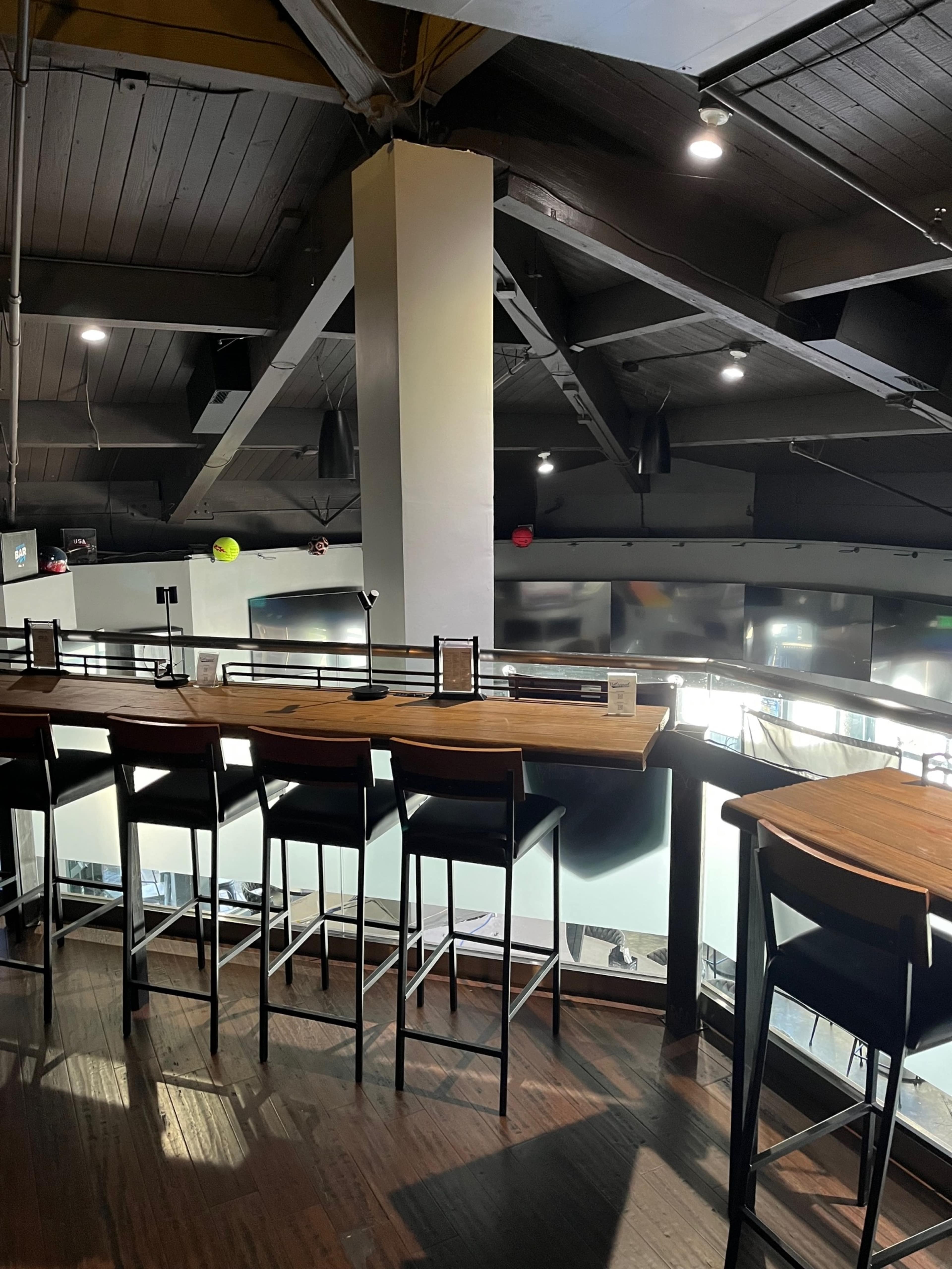 The image shows a modern, spacious interior of a bar or restaurant with high tables and tall stools, featuring open wooden beams and a view of a lower level.