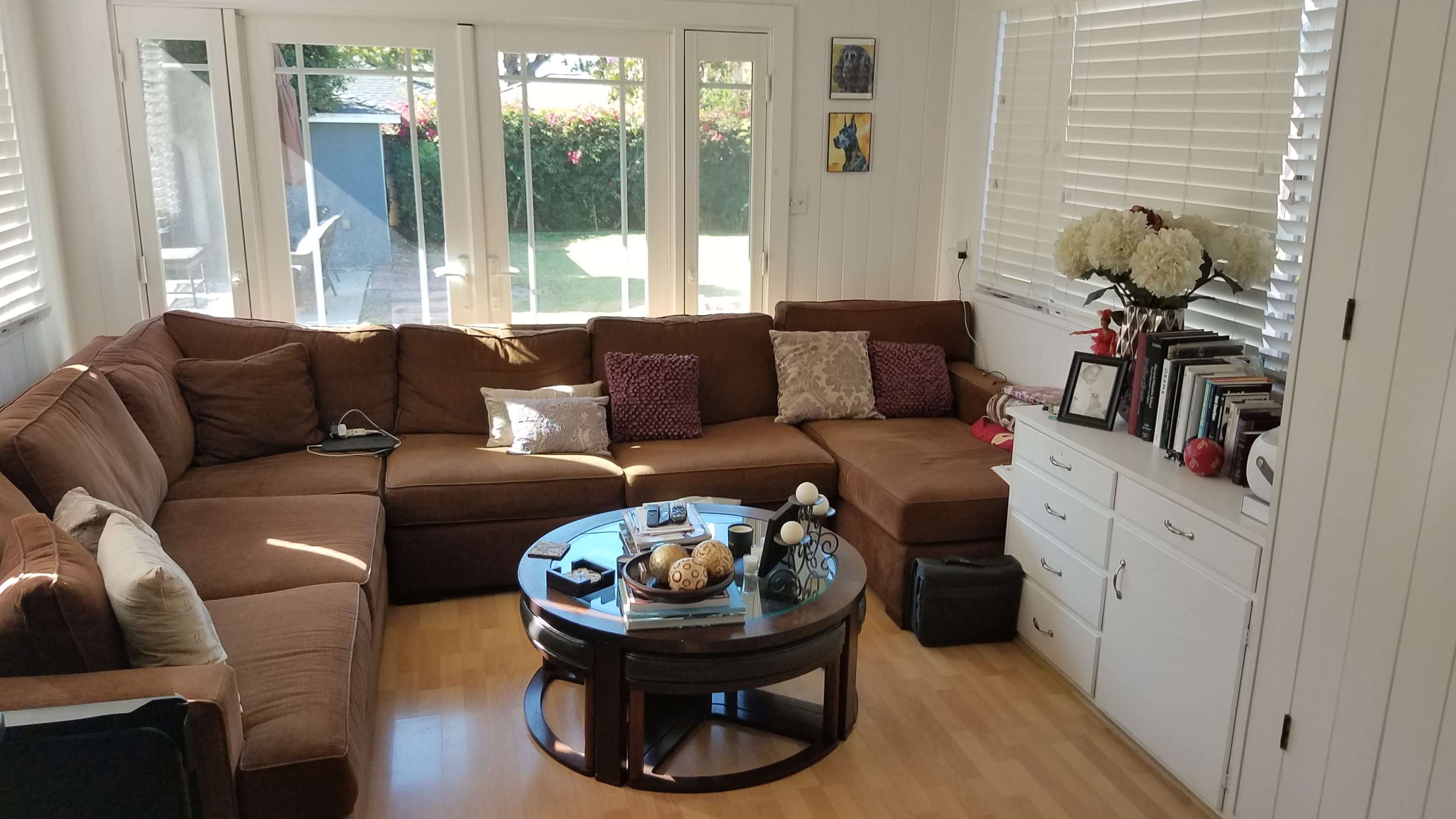 The image shows a brown sectional sofa arranged around a round coffee table in a sunlit living room with white walls and large windows.