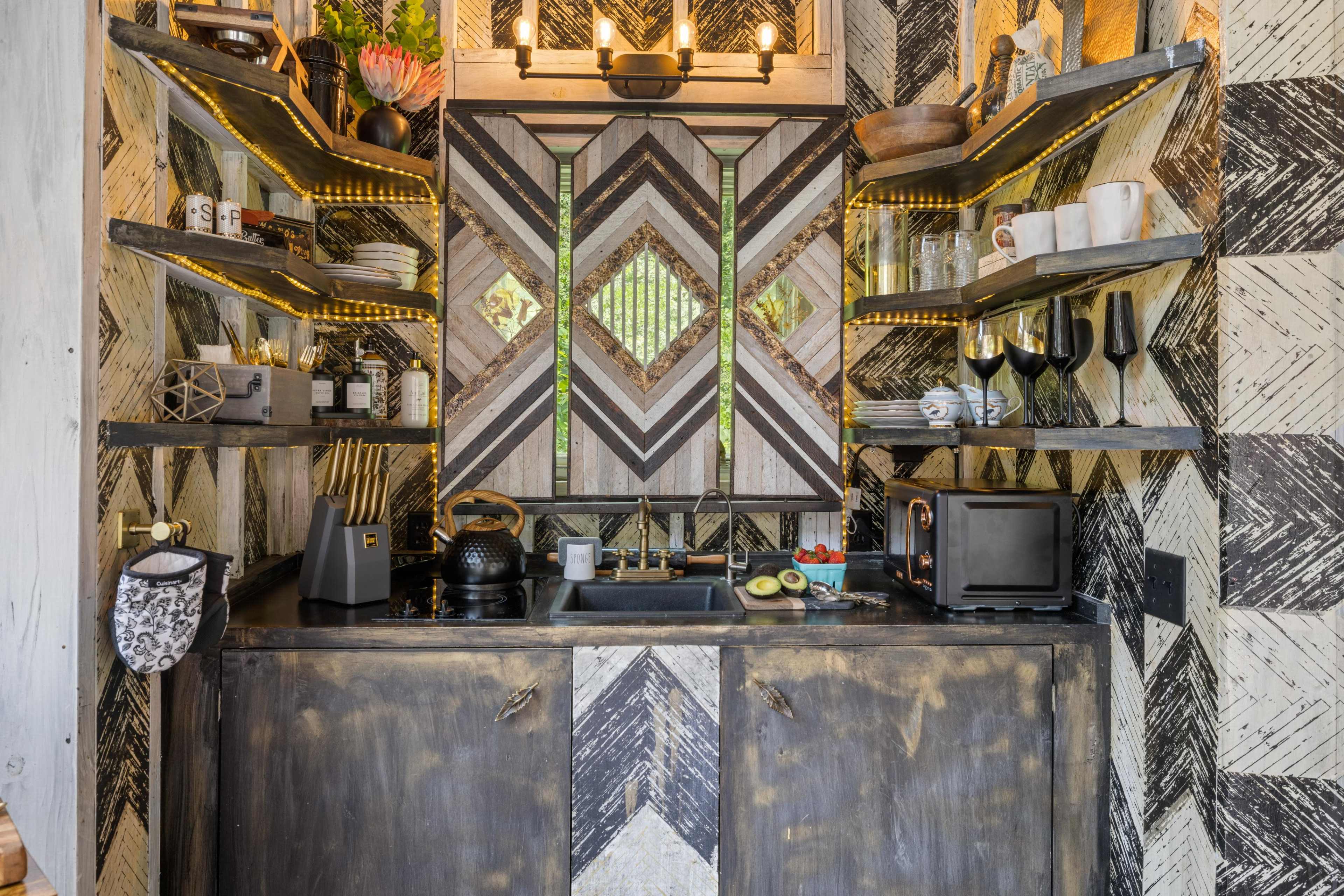 A compact kitchen area featuring a dark wood cabinetry with geometric-patterned walls, shelves holding various dishes and decor, and a small sink with appliances and fresh produce on the counter.