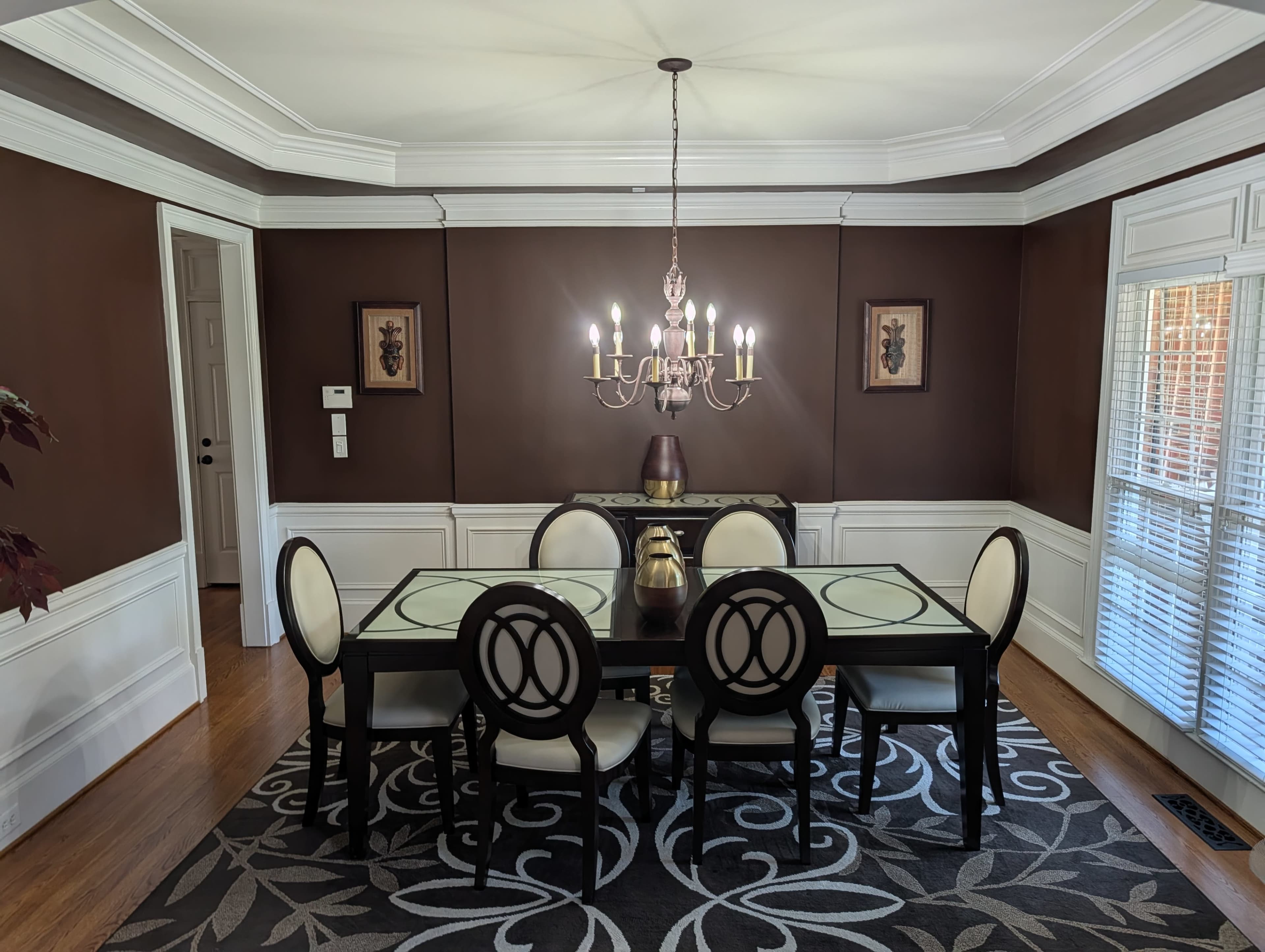 The image shows a dining room with a rectangular glass table surrounded by upholstered chairs, a chandelier hanging from the ceiling, and dark brown walls adorned with decorative art.