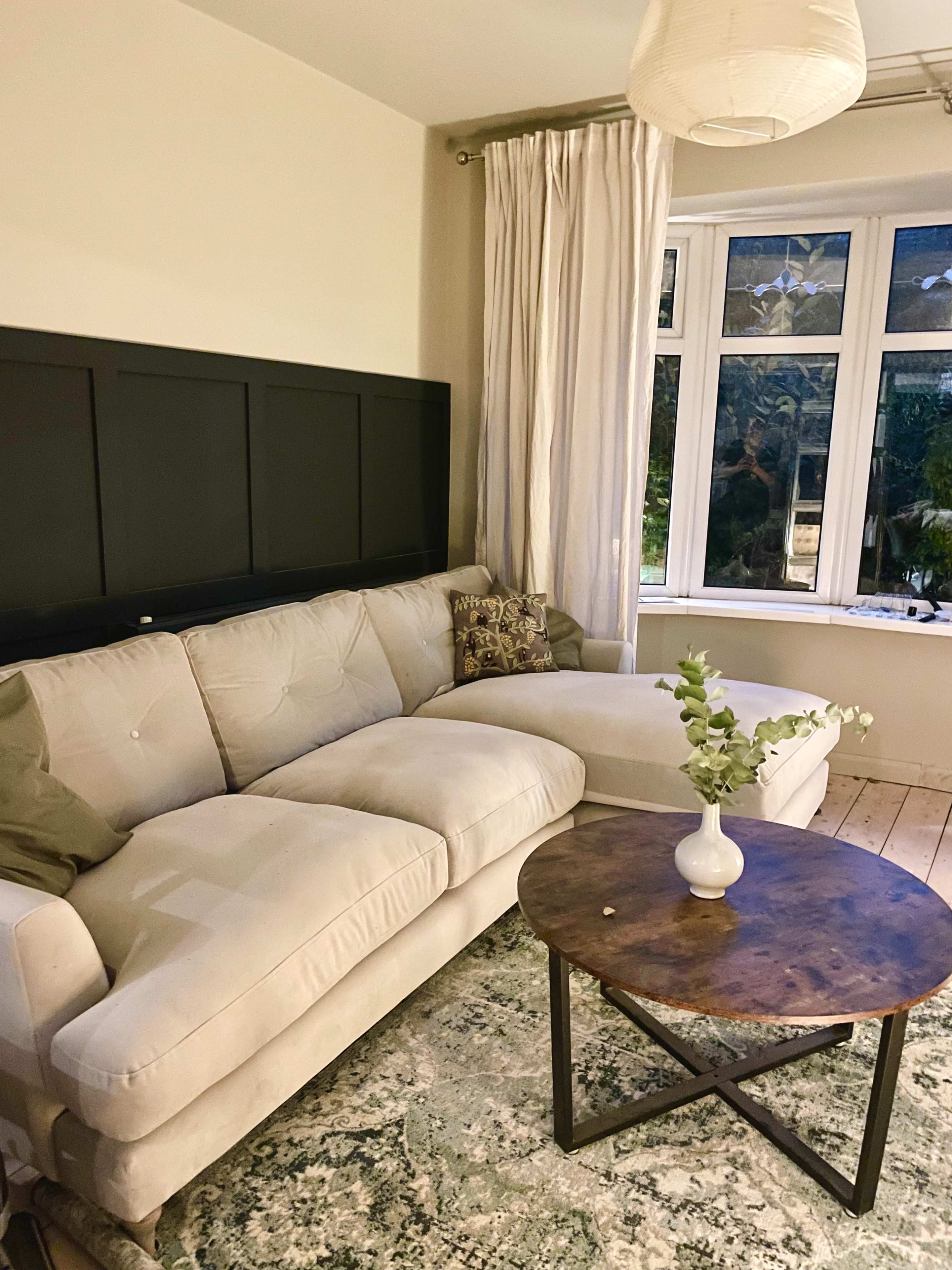 A light-colored sectional sofa with a decorative cushion is positioned next to a round wooden coffee table in a room with a large window and sheer curtains.