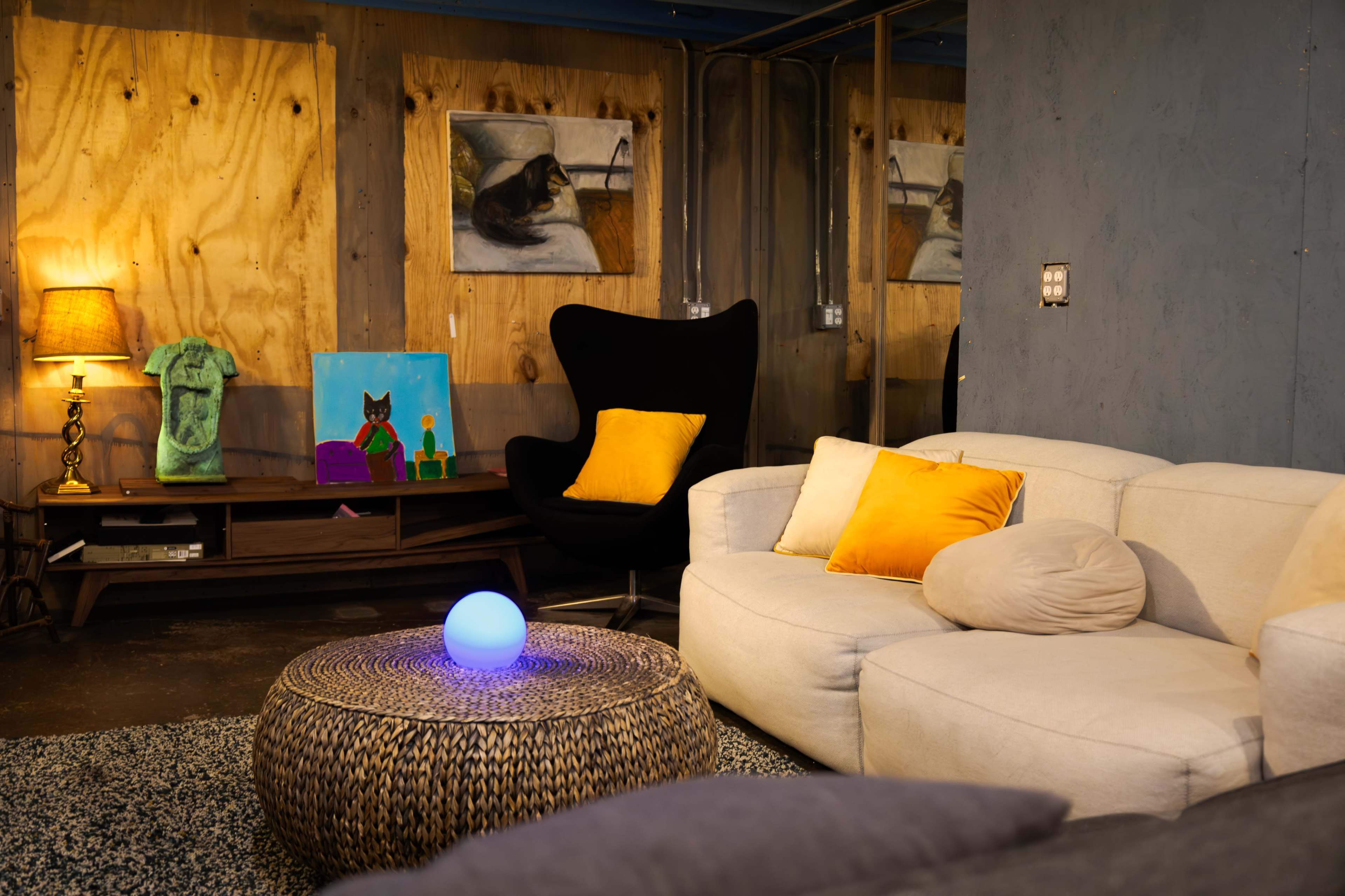 The image features a modern living room with a white sofa, a round woven coffee table, and colorful artwork on the walls, including a painting of a cat.