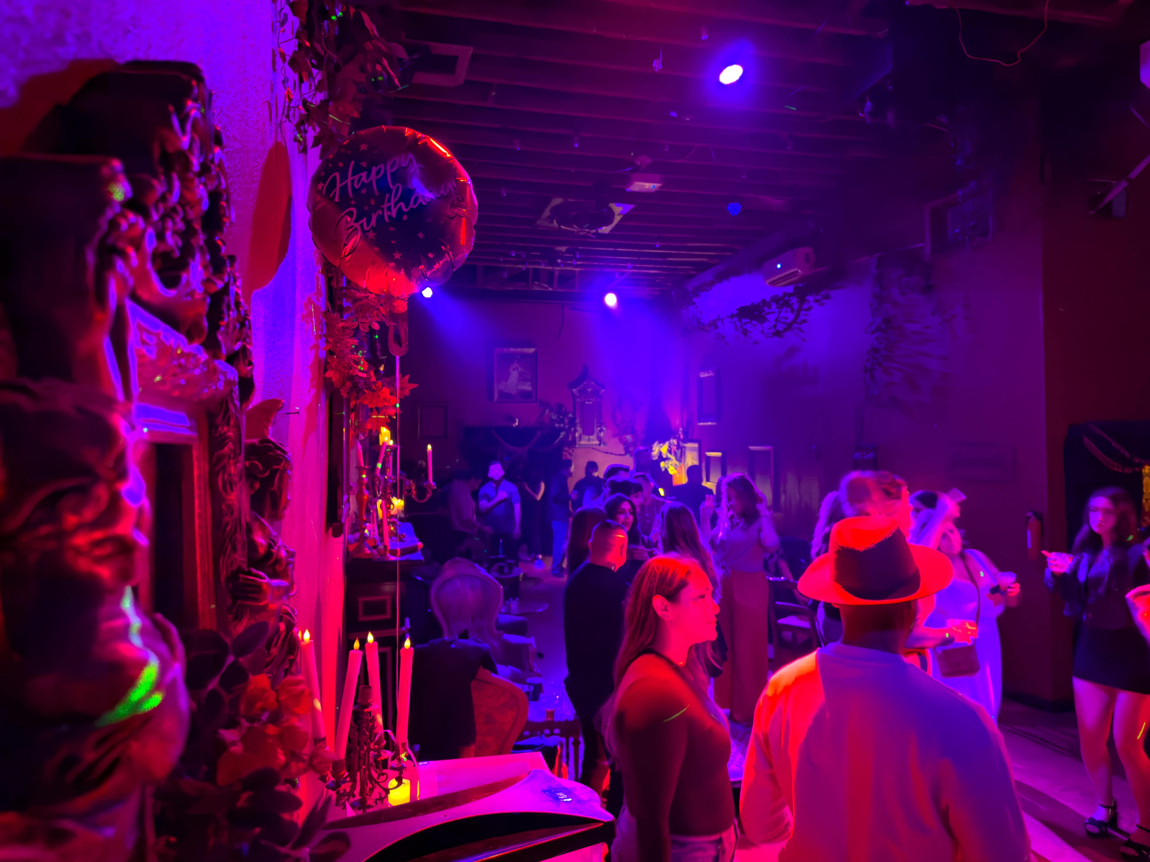 A vibrant party scene filled with people dancing under colorful lights, featuring decorations and a "Happy Birthday" balloon.
