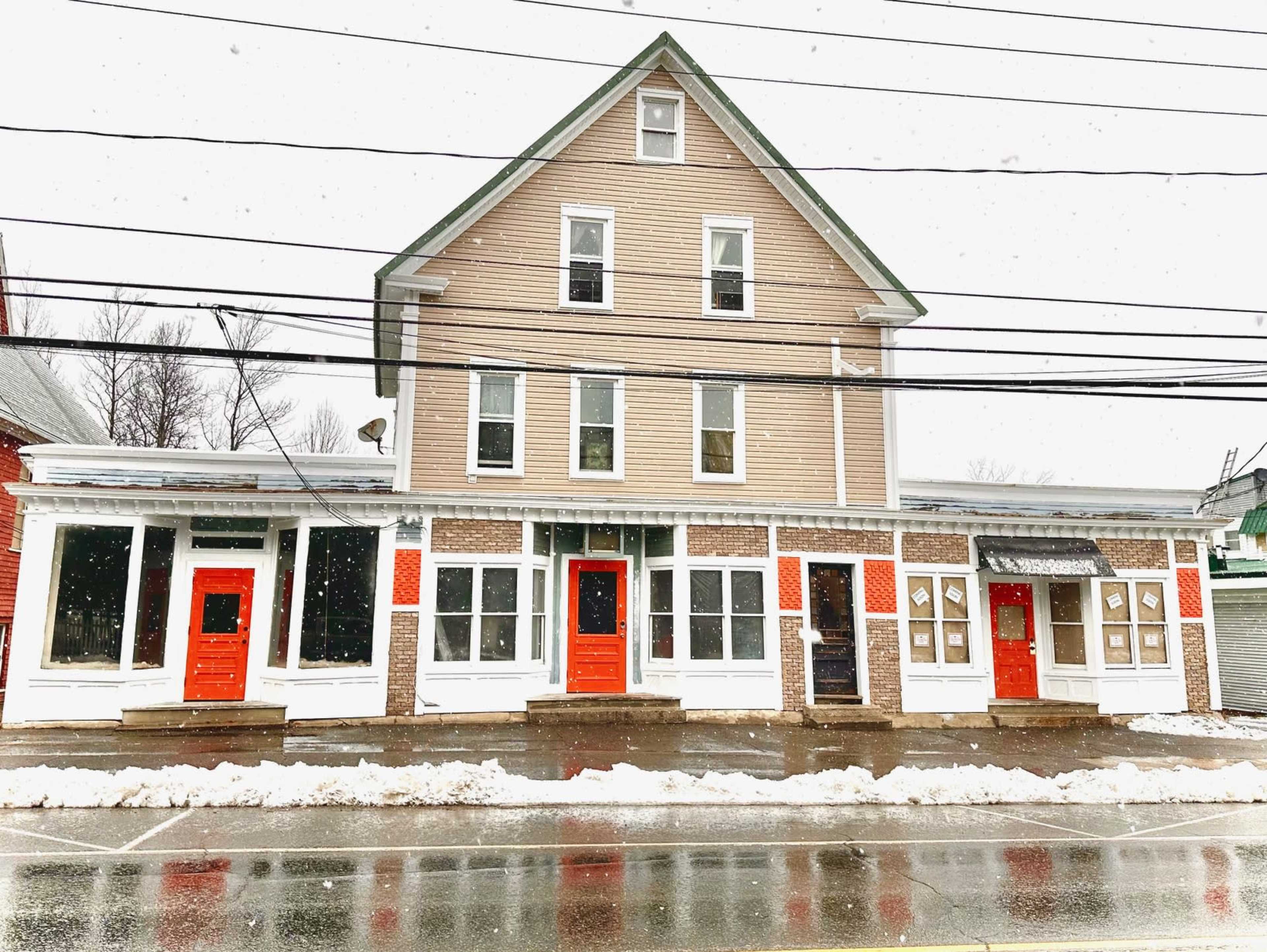 A three-story building with a light brown exterior and red doors is seen on a snowy street, reflecting in the wet pavement.
