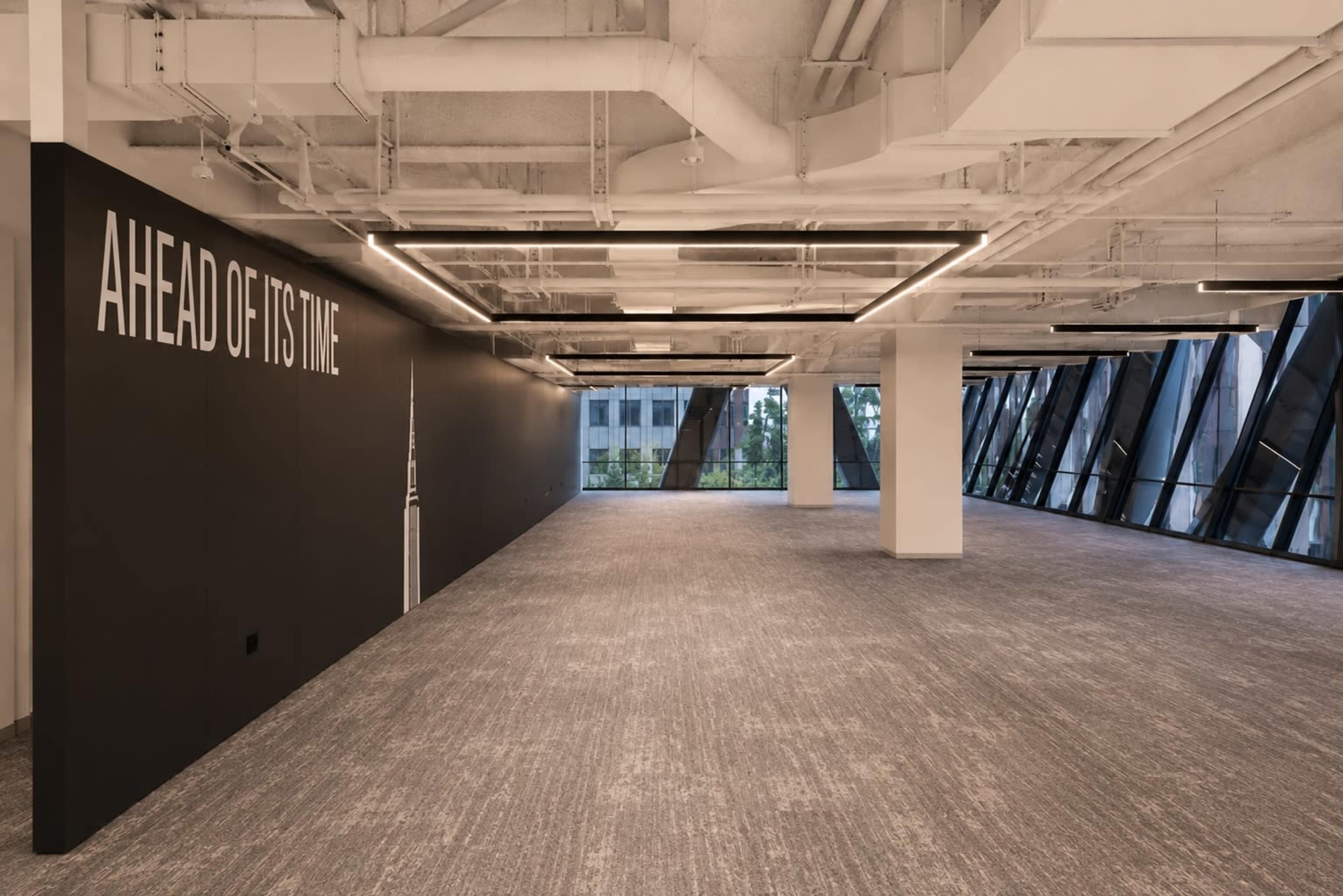 The image shows an empty office space with a black wall featuring the phrase "AHEAD OF ITS TIME" and large windows displaying a cityscape.
