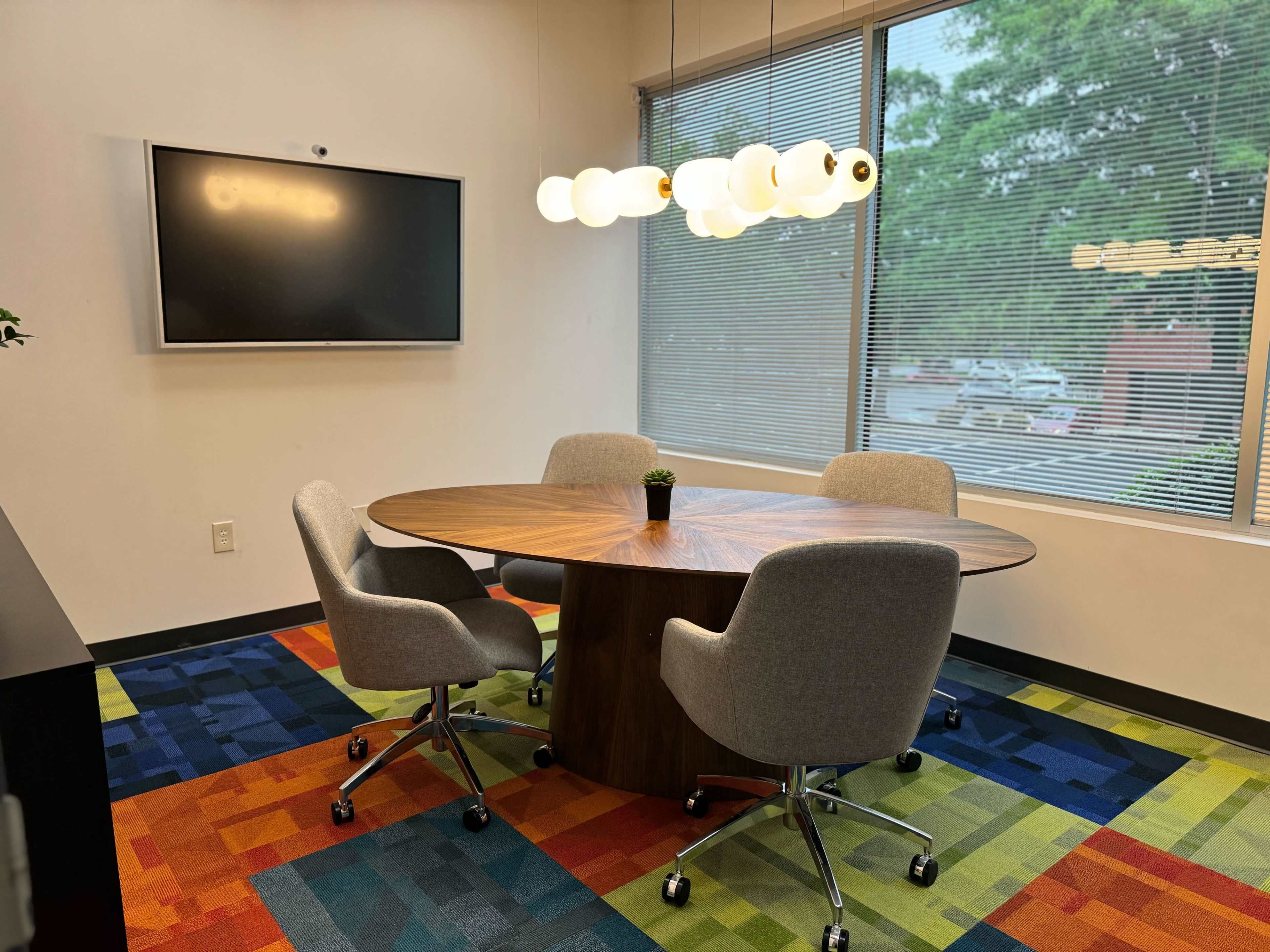 A modern conference room features a round wooden table surrounded by four gray chairs, with a large screen on the wall and a colorful carpet.