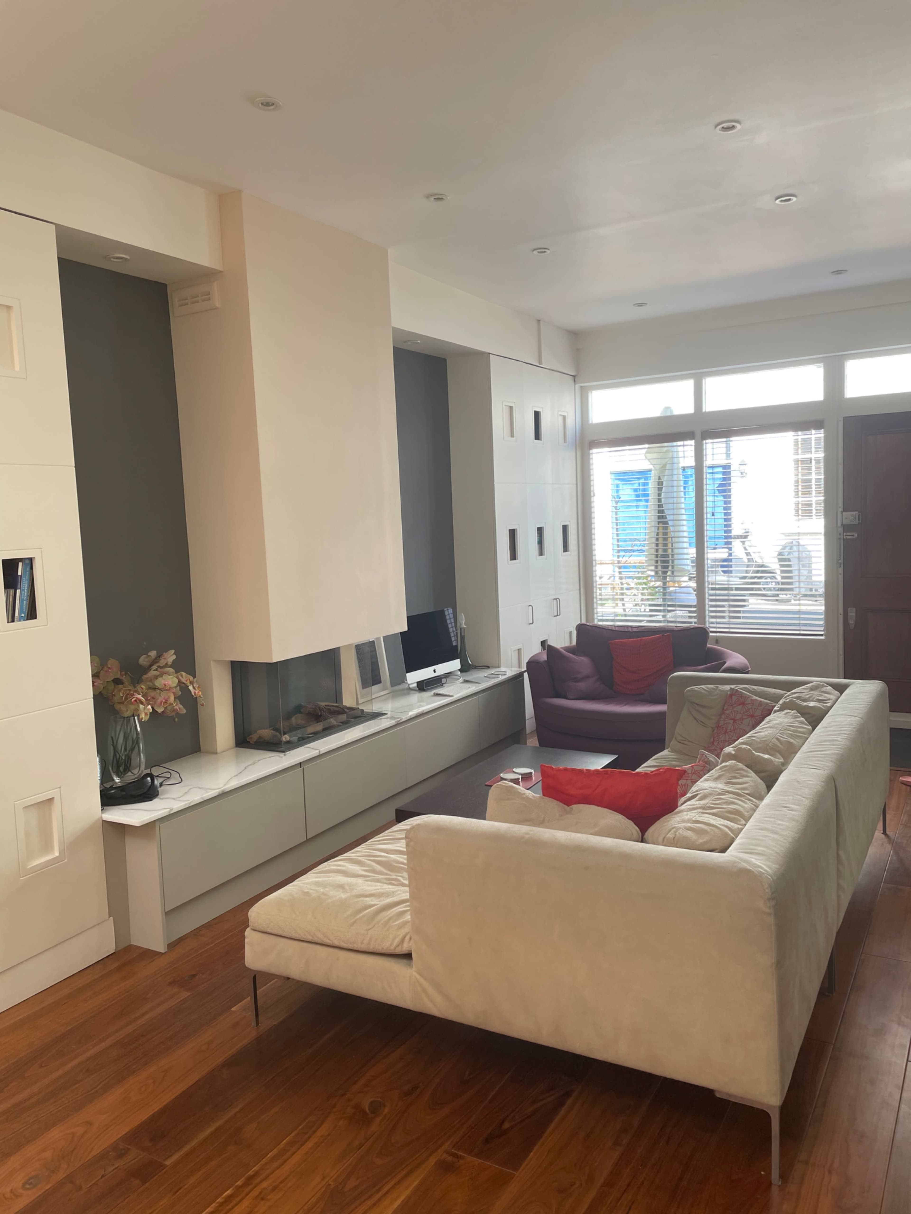 A modern living room with a light-colored sectional sofa, a wall-mounted fireplace, and large windows allowing natural light to enter.
