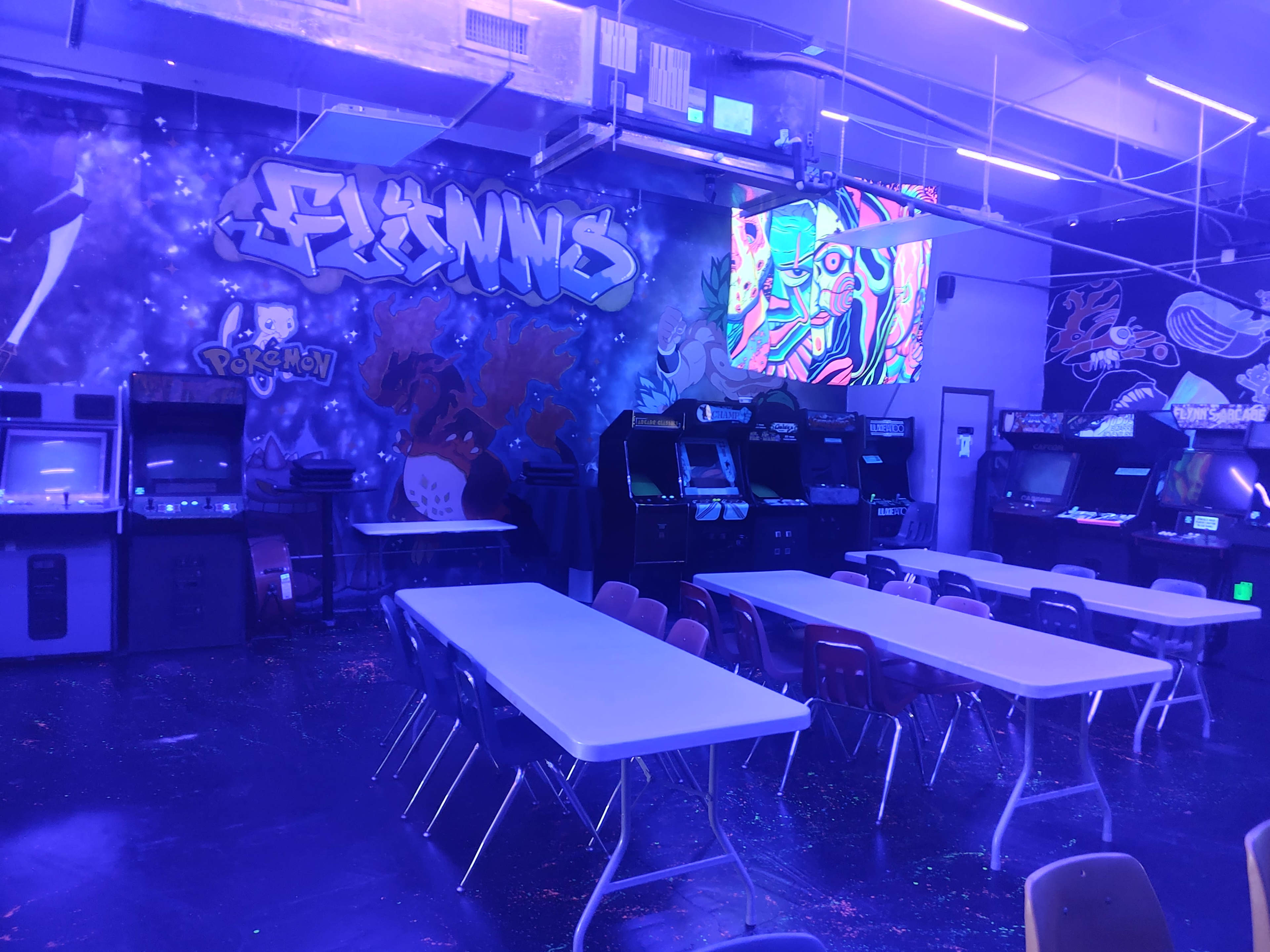 The image shows a gaming arcade with arcade machines, tables, and a colorful mural on the wall.