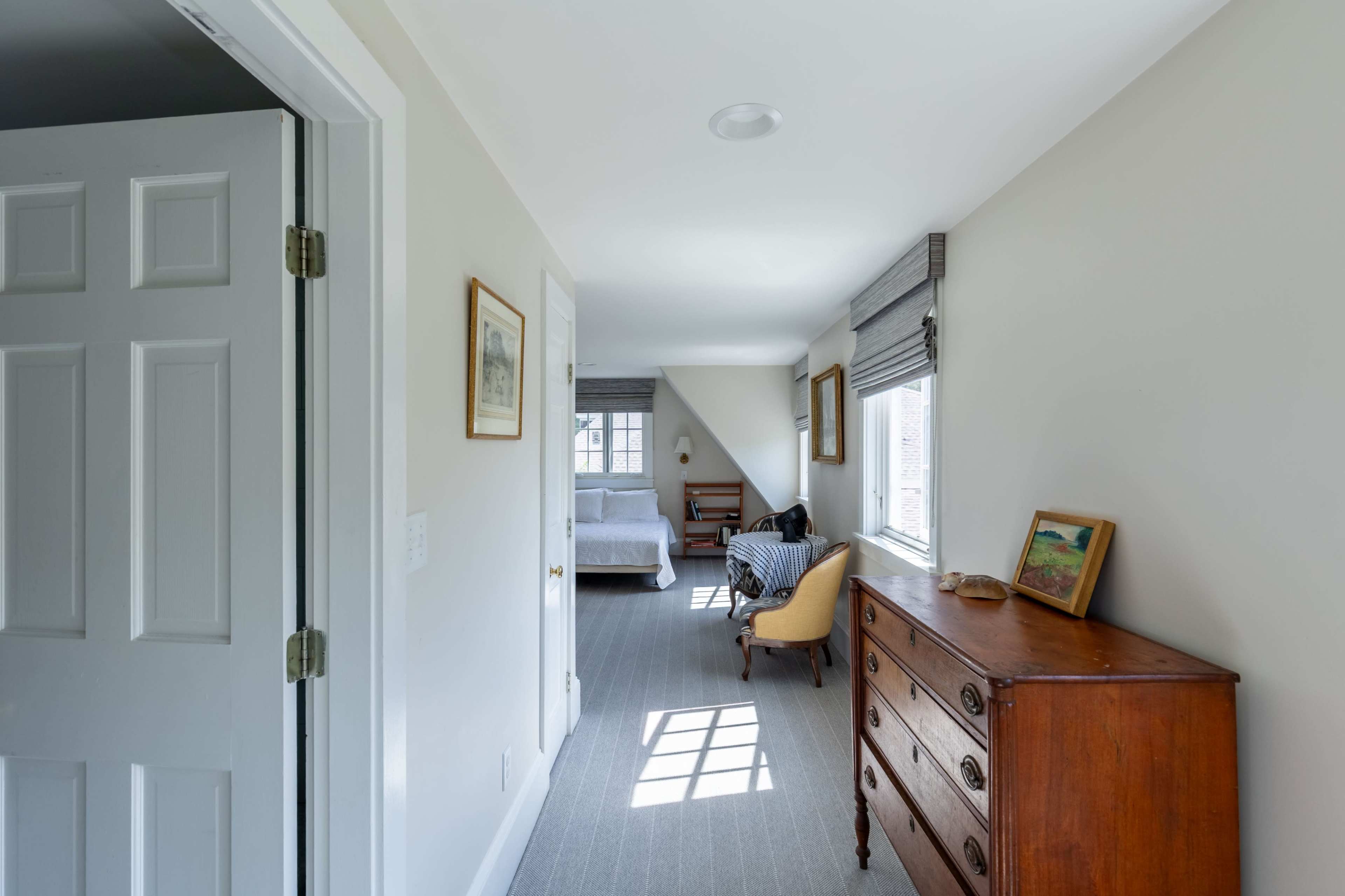 A bright hallway leads to a bedroom, featuring a wooden dresser and a chair beside a window.