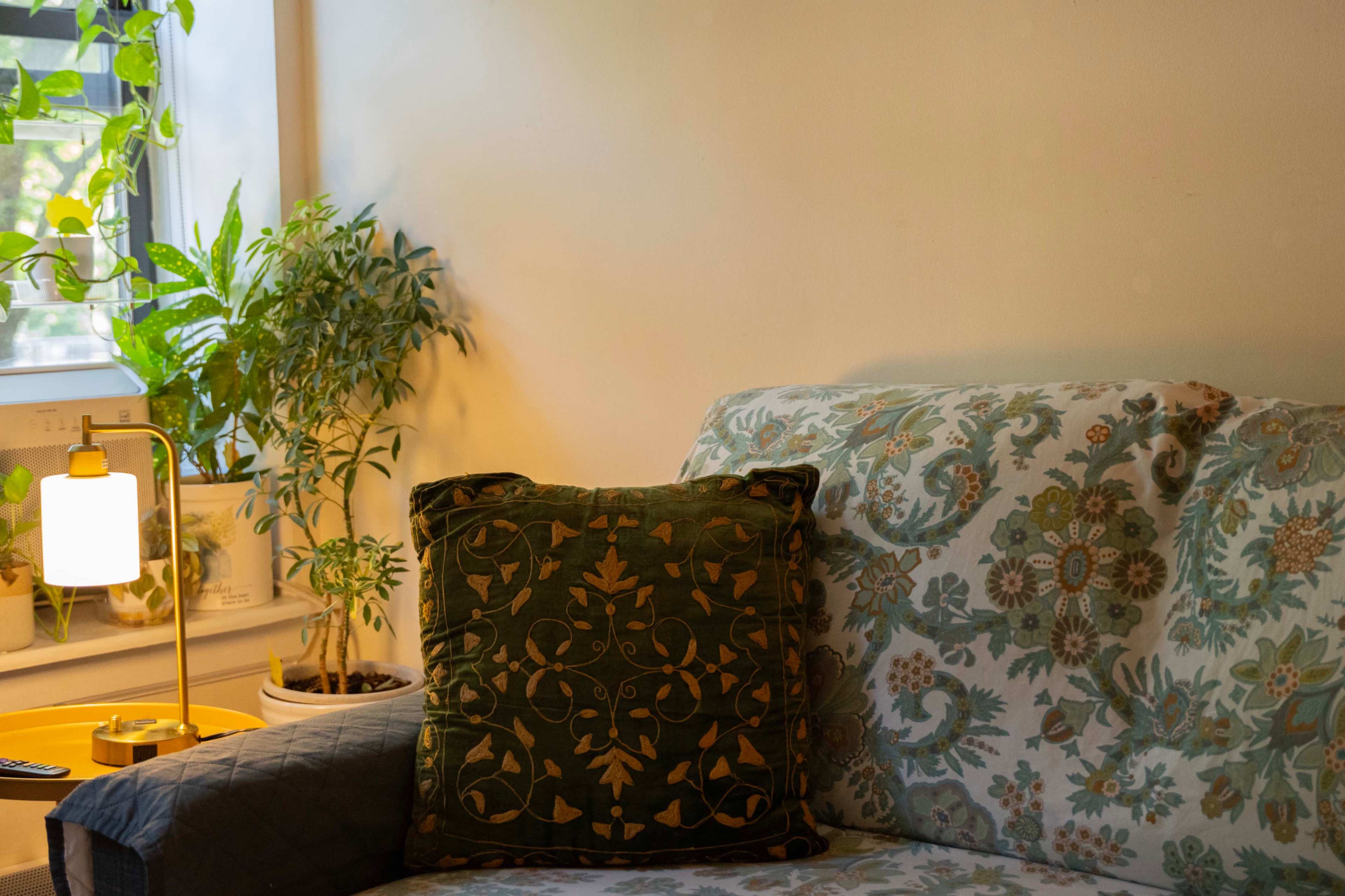 A sofa with a patterned throw pillow is positioned next to a small lamp and a potted plant in a well-lit room.