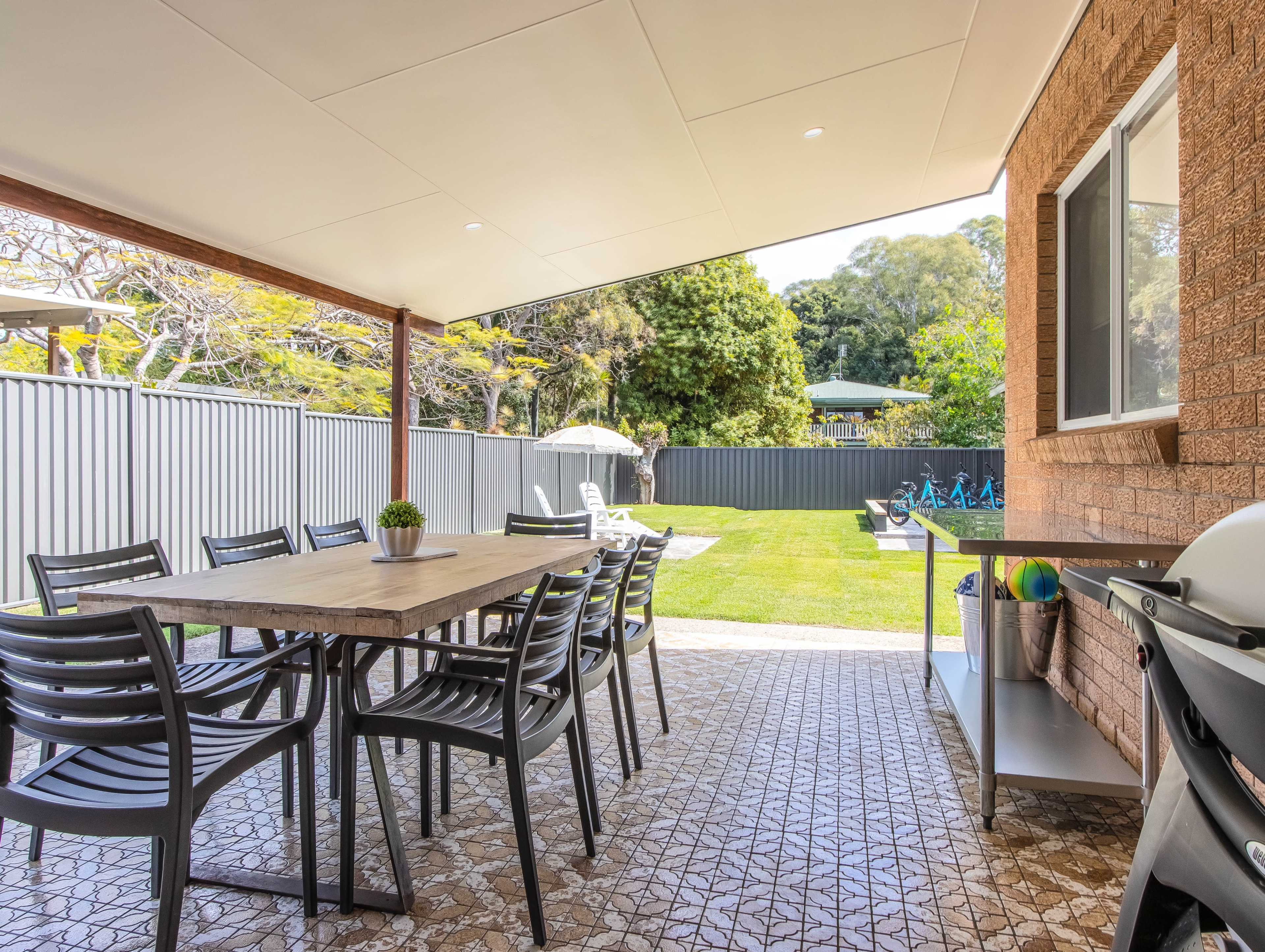A covered outdoor dining area features a large table surrounded by chairs, with a view of a landscaped backyard and a grill nearby.