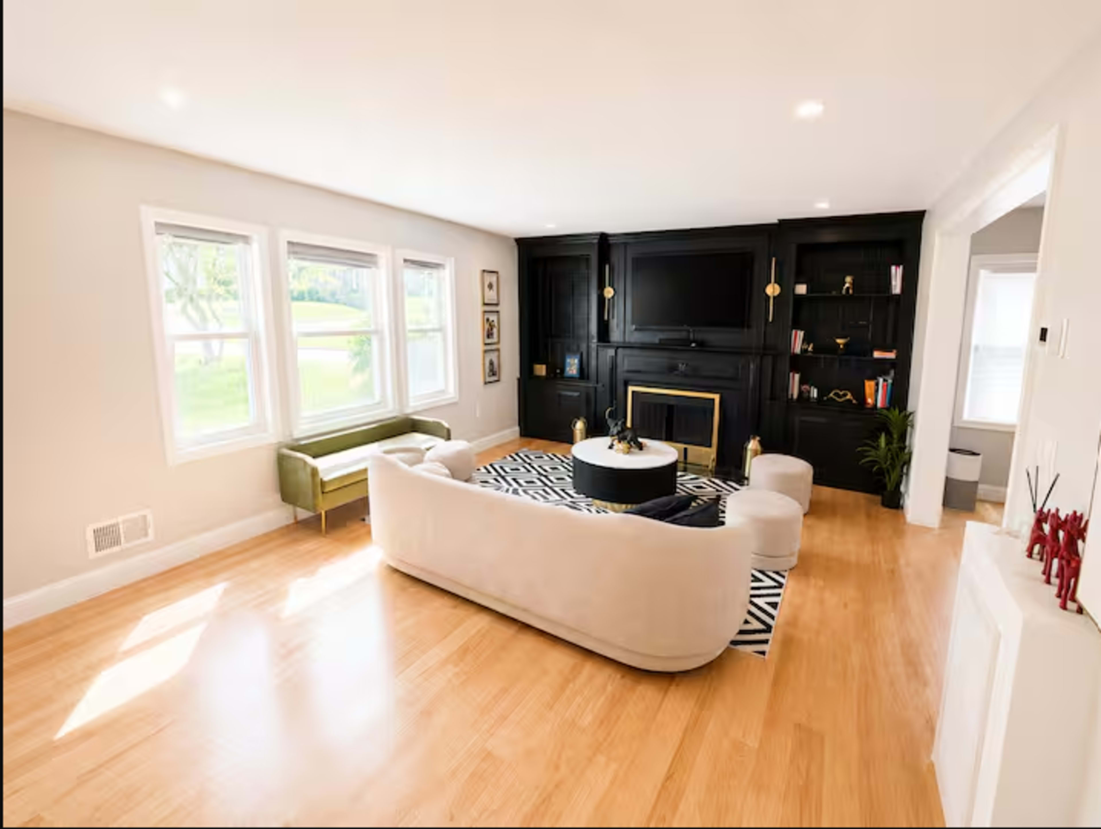 The living room features a light-colored sofa and armchairs, a round coffee table, a black entertainment center, and large windows that allow natural light to fill the space.