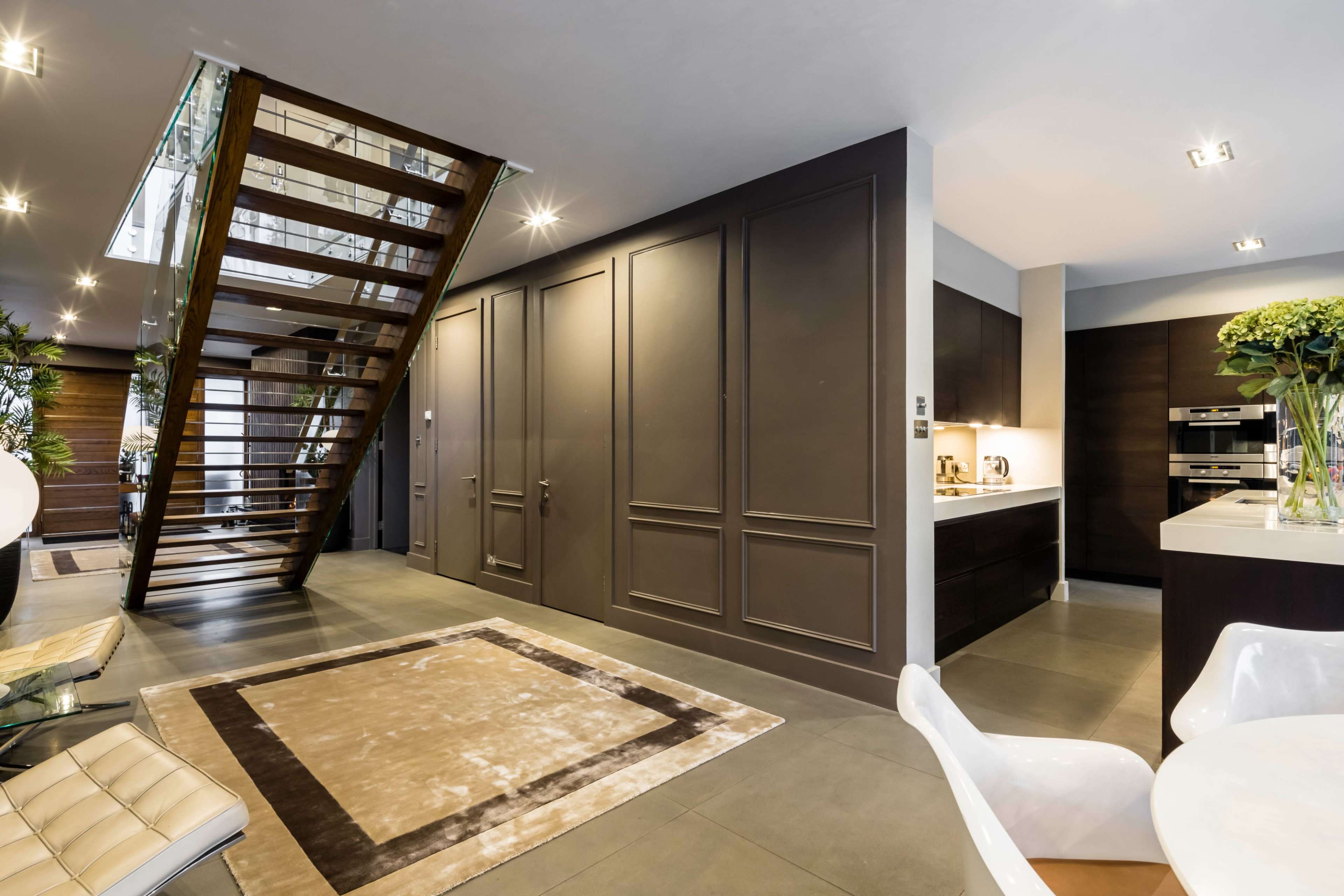 The image shows a modern interior featuring a staircase, a kitchen area, and a dining space with minimalist furniture and decorative plants.