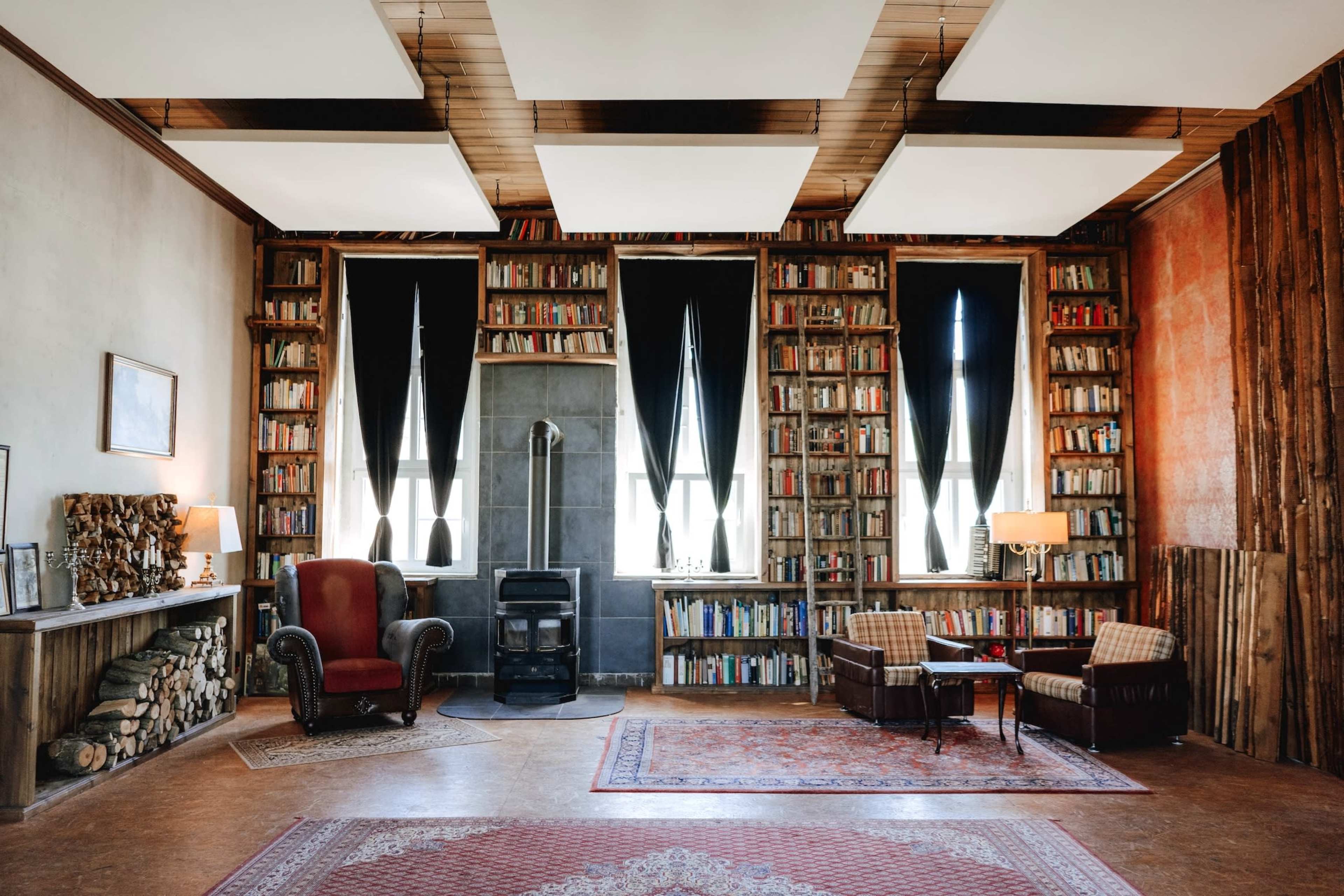 The image depicts a cozy library with wooden shelves filled with books, two armchairs, a small table, and a wood-burning stove.