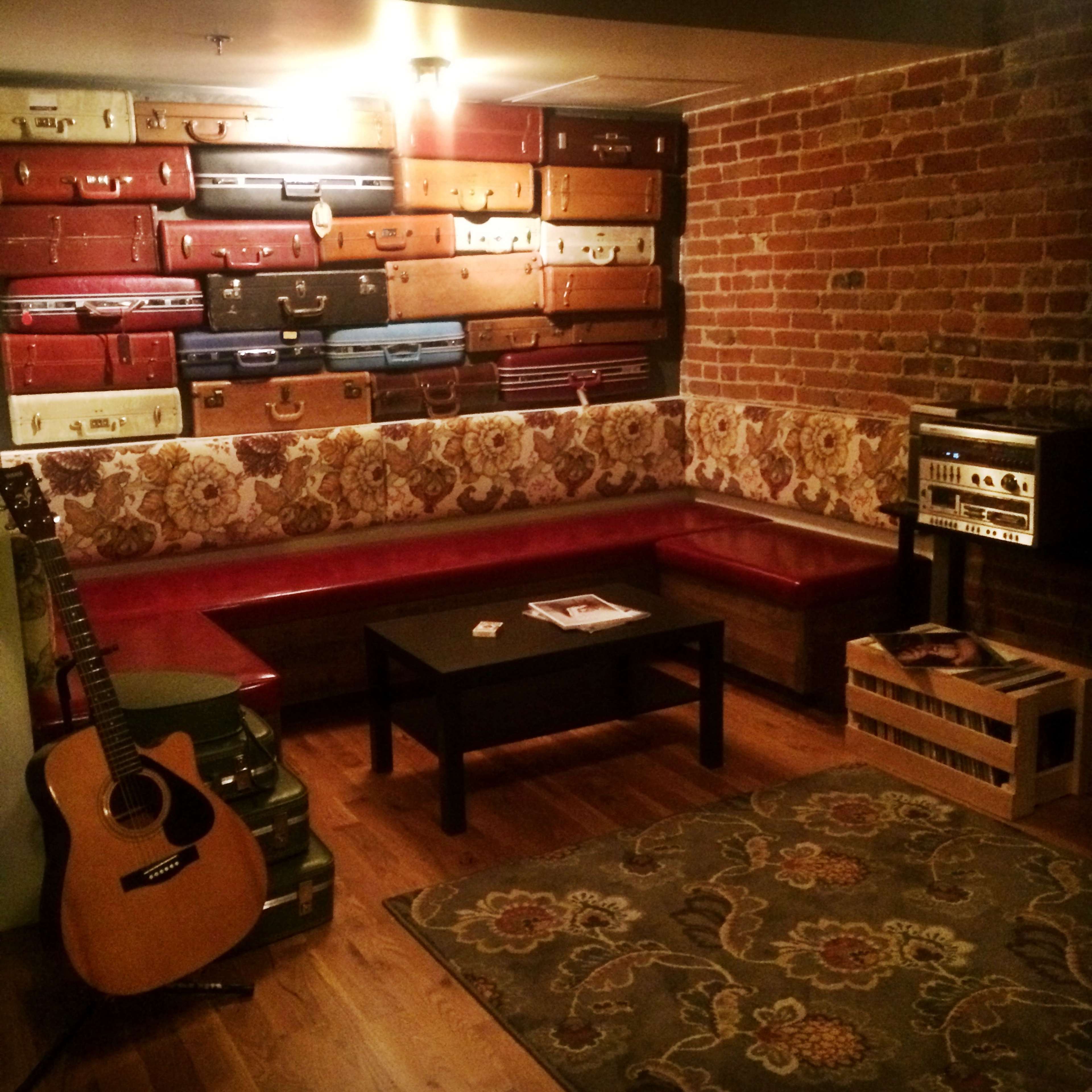 A cozy lounge area features a wall adorned with vintage suitcases, a wooden table, a guitar, and a patterned rug.
