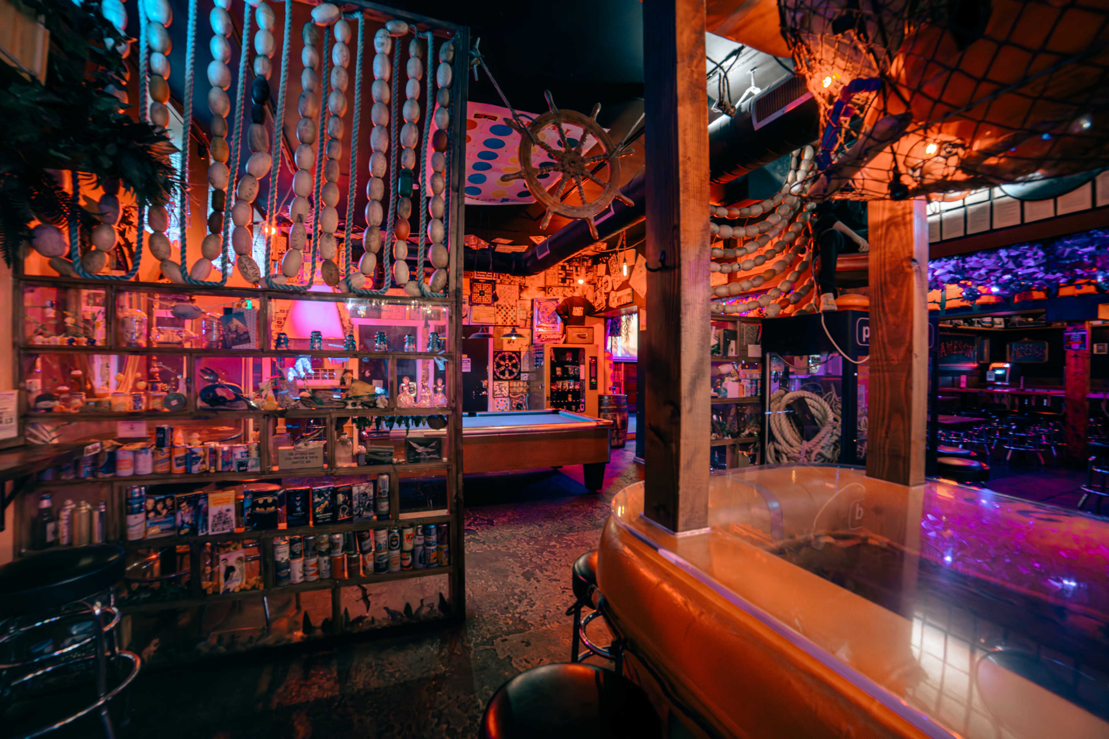 The interior of a bar features colorful lighting, a pool table, and shelves filled with various bottles and decorations, creating a lively atmosphere.