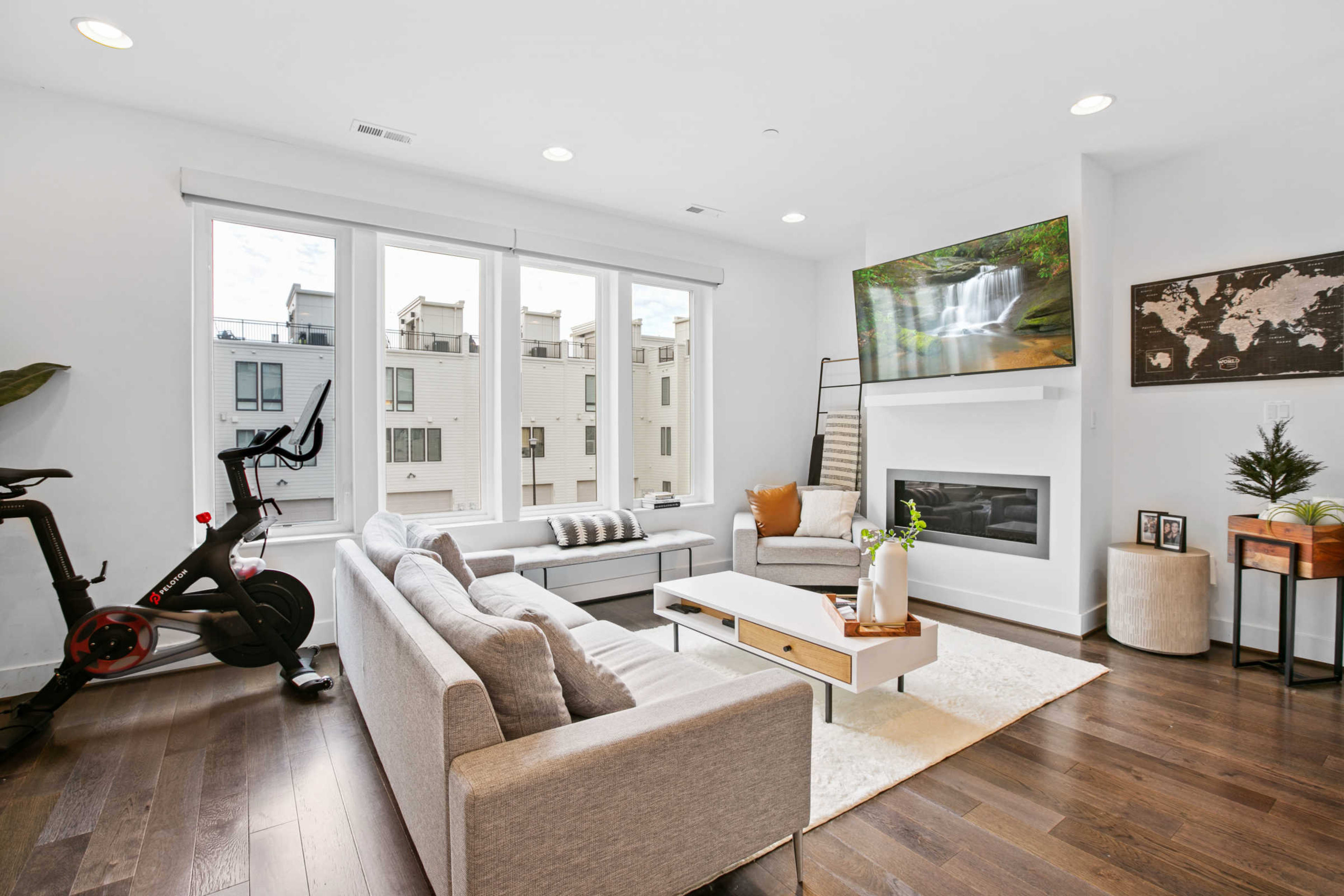 The living room features a modern design with large windows, a fireplace, a mounted TV, and a workout bike in the corner.