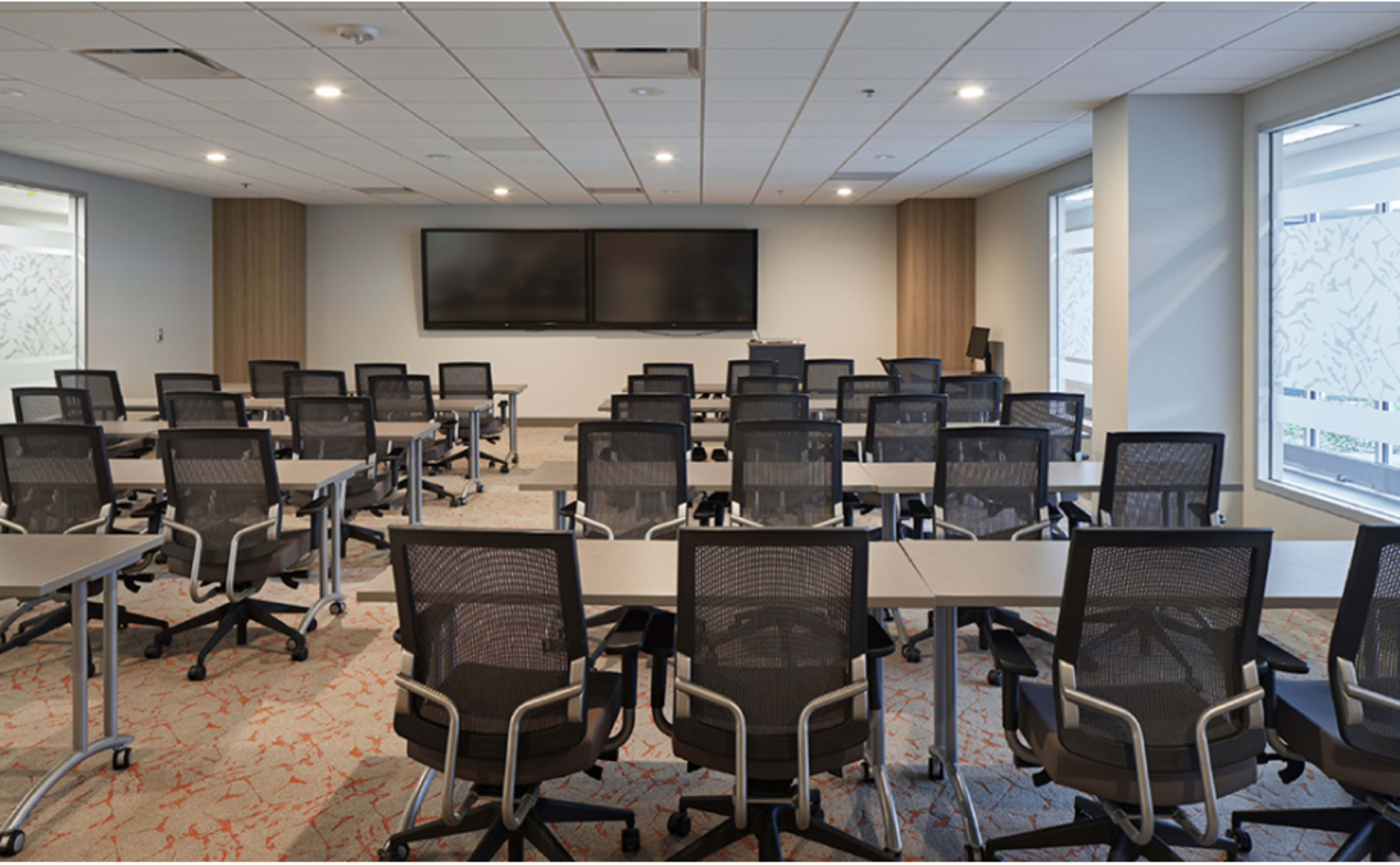 A modern conference room arranged with multiple rows of desks and ergonomic chairs facing two large screens on the front wall.