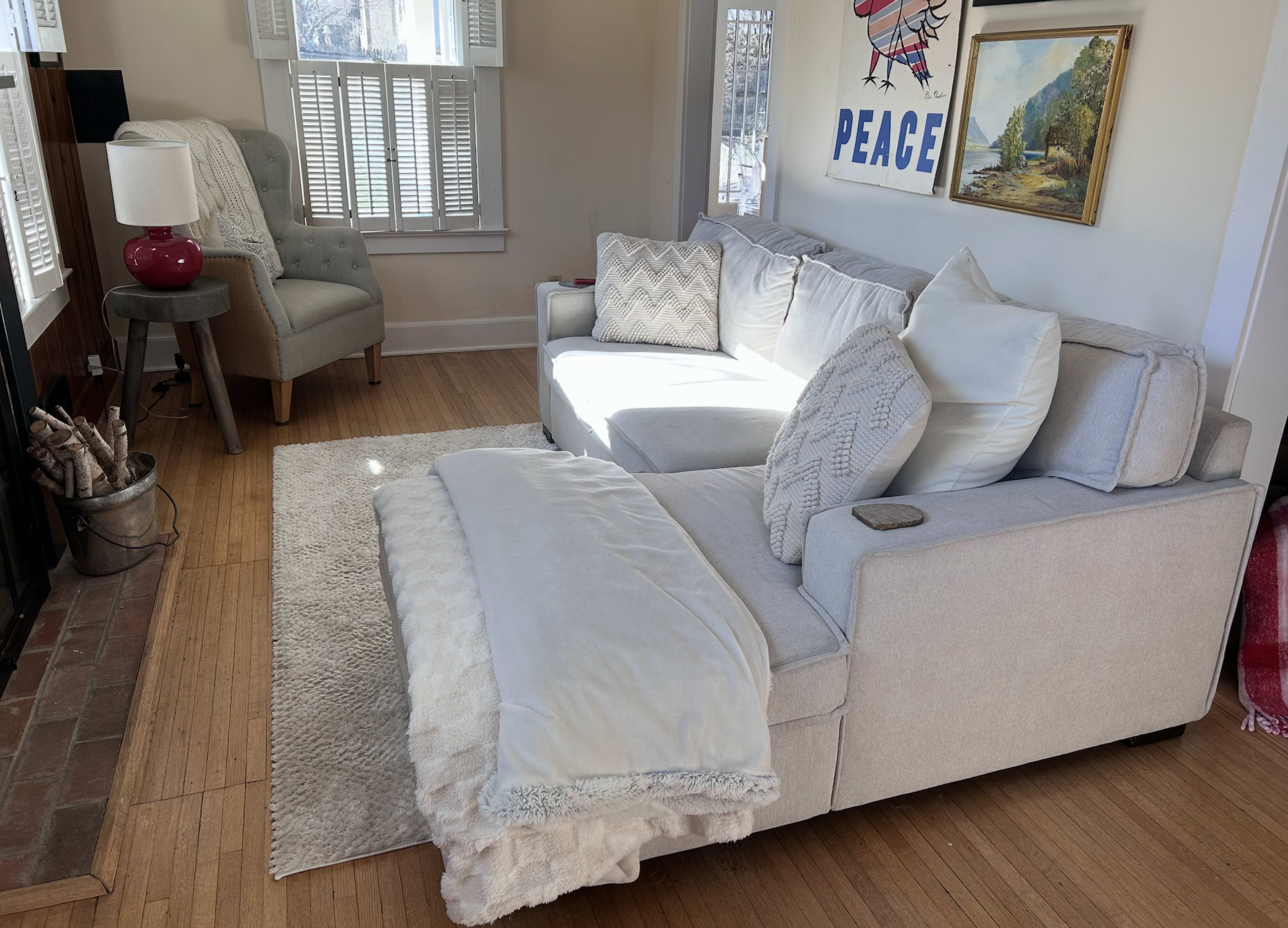 The image shows a cozy living room with a light-colored sectional sofa, a small table with a lamp, and a chair near a window, all arranged on a bamboo floor.