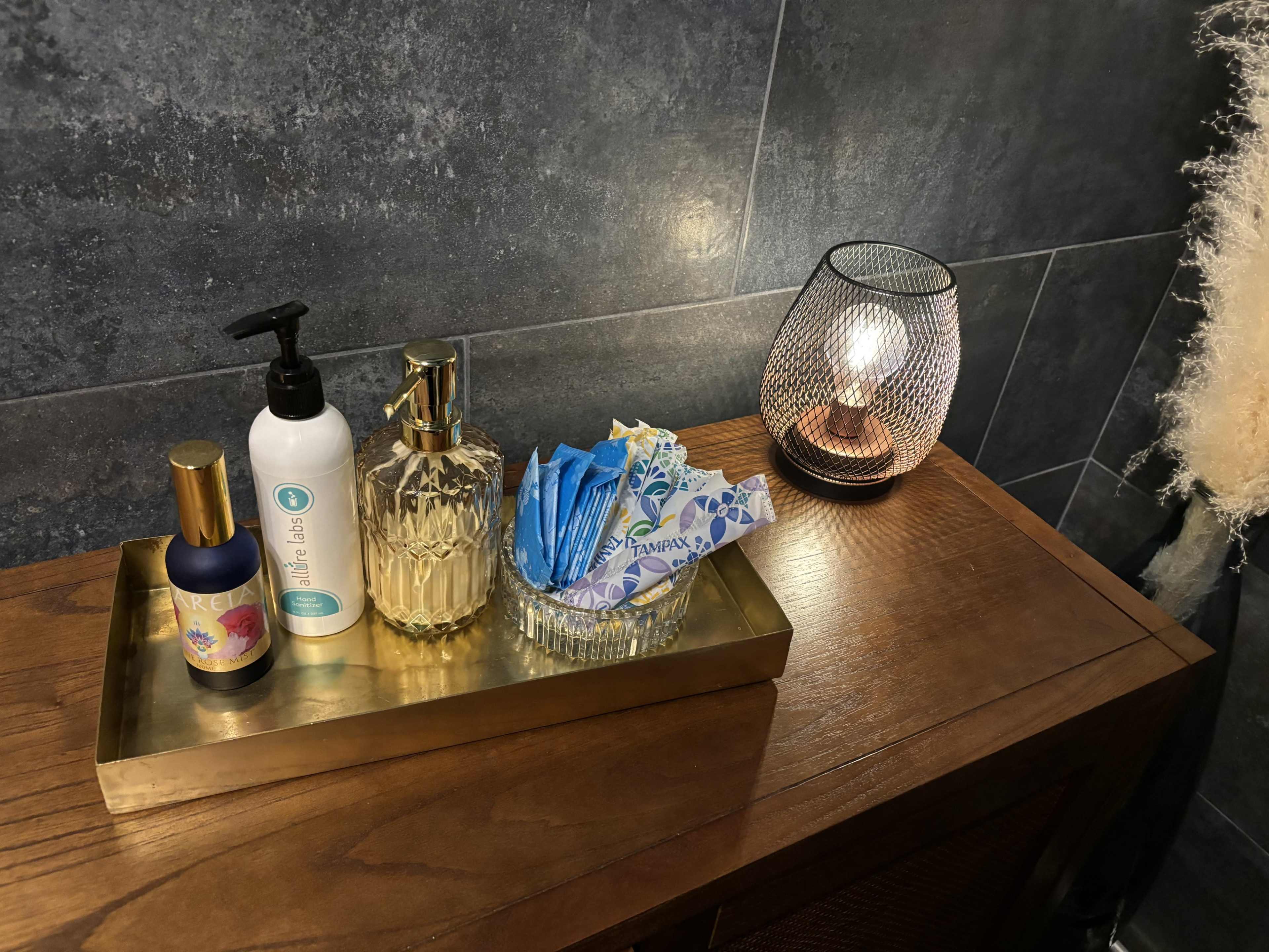 A wooden tray holds various bathroom toiletries including a spray bottle, lotion, and items wrapped in blue paper, beside a decorative lamp.