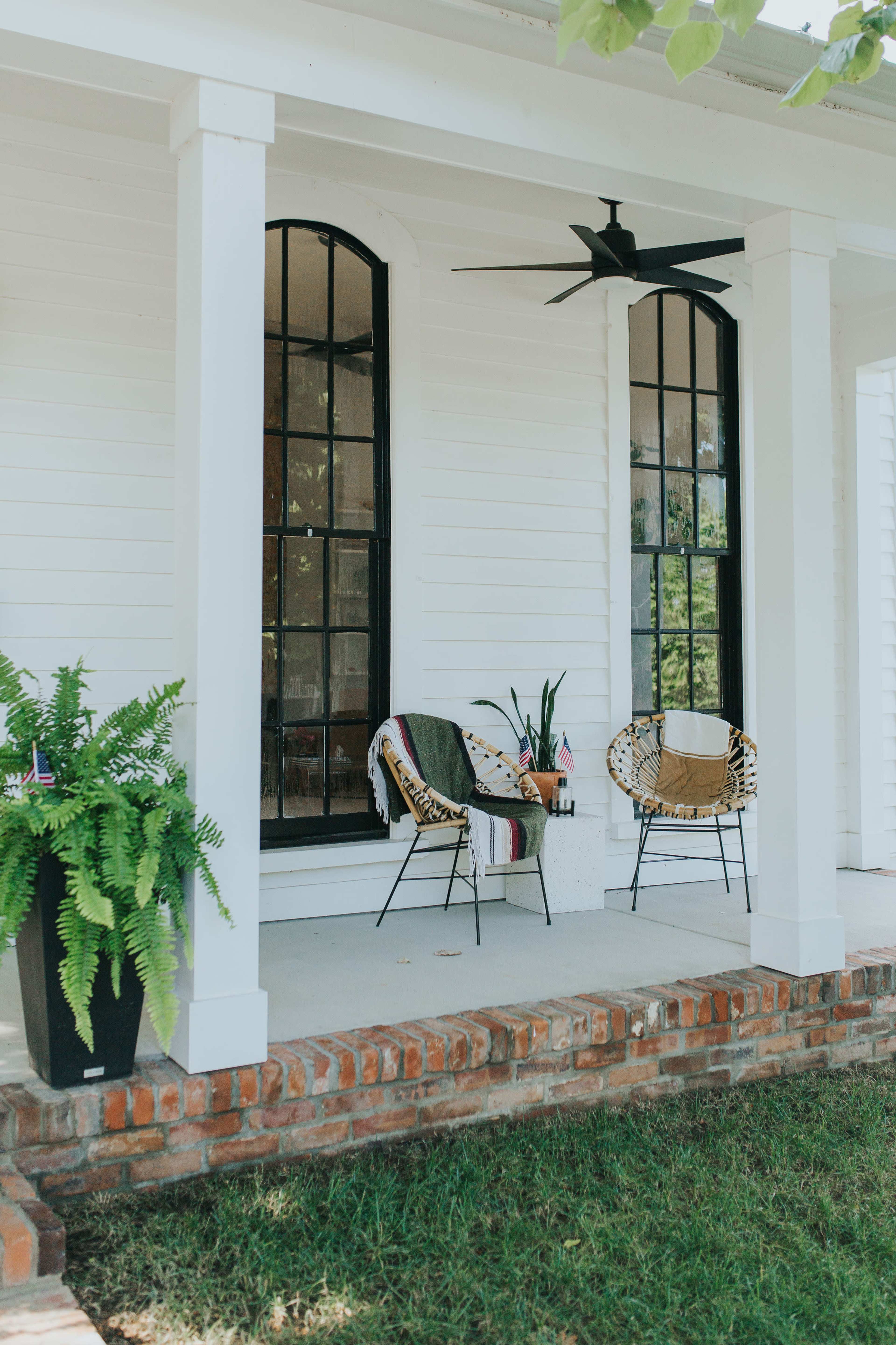 A covered porch featuring two chairs, a potted plant, and a small table, with a brick base and large windows in the background.