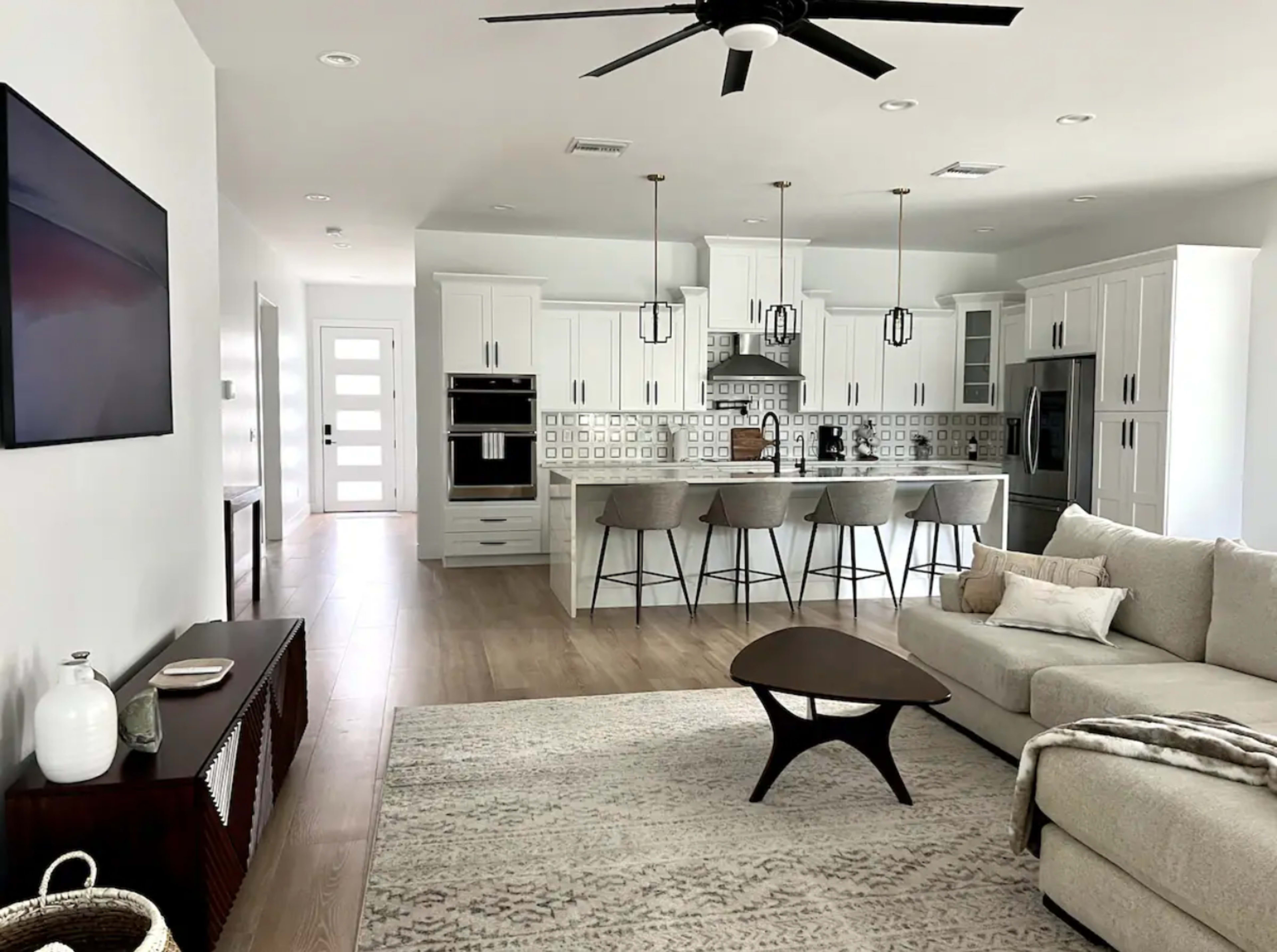 A modern living room features a light-colored sofa, a coffee table, and a view of a kitchen with white cabinets and pendant lights.