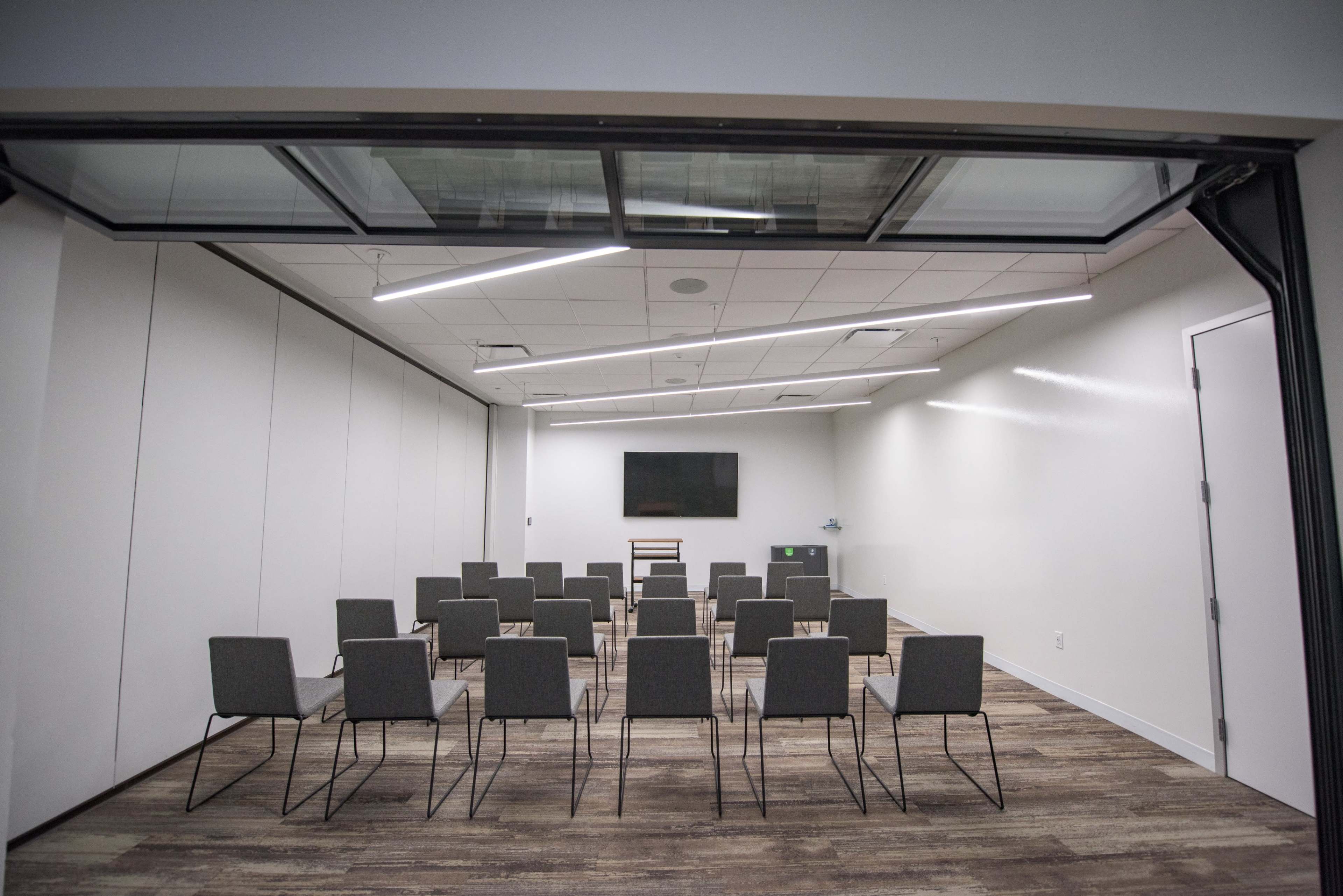 A modern conference room features rows of gray chairs facing a wall-mounted screen.