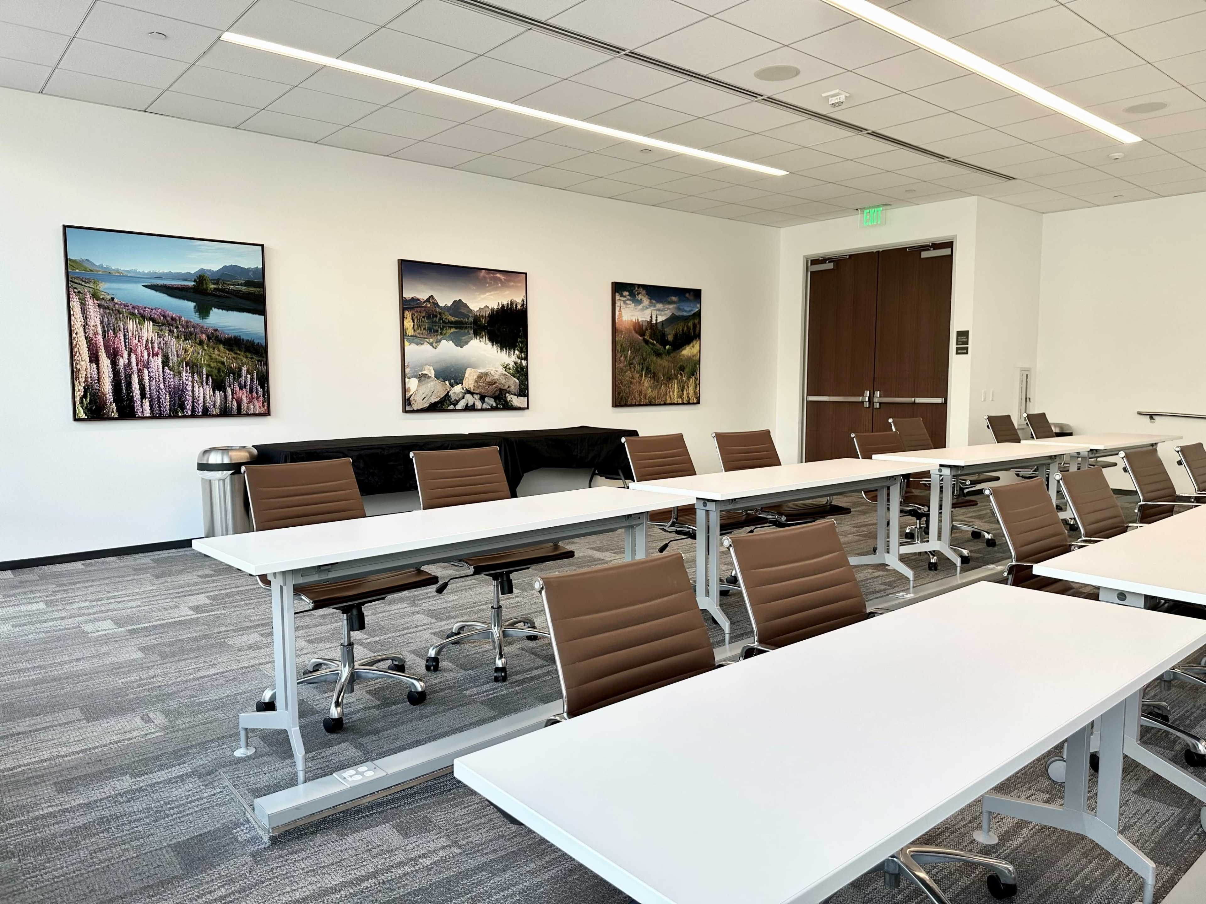 A modern conference room features several tables and chairs arranged for a meeting, with landscape artwork hanging on the walls.