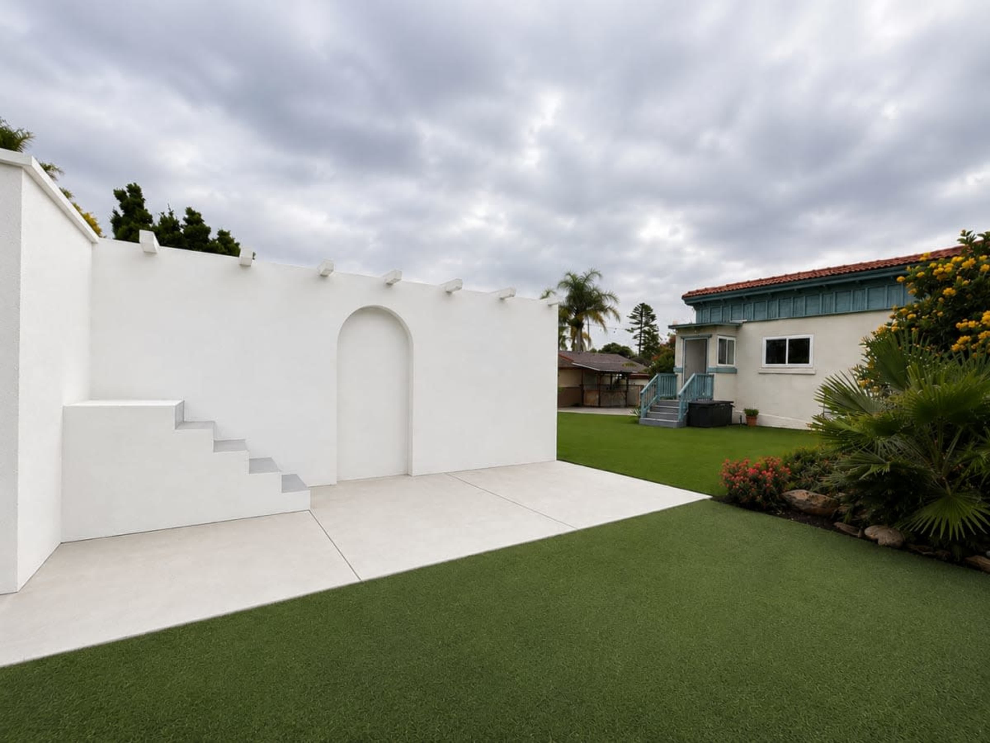 The image shows a spacious backyard with a white wall featuring an arched opening, artificial turf, and a two-story residential building in the background.