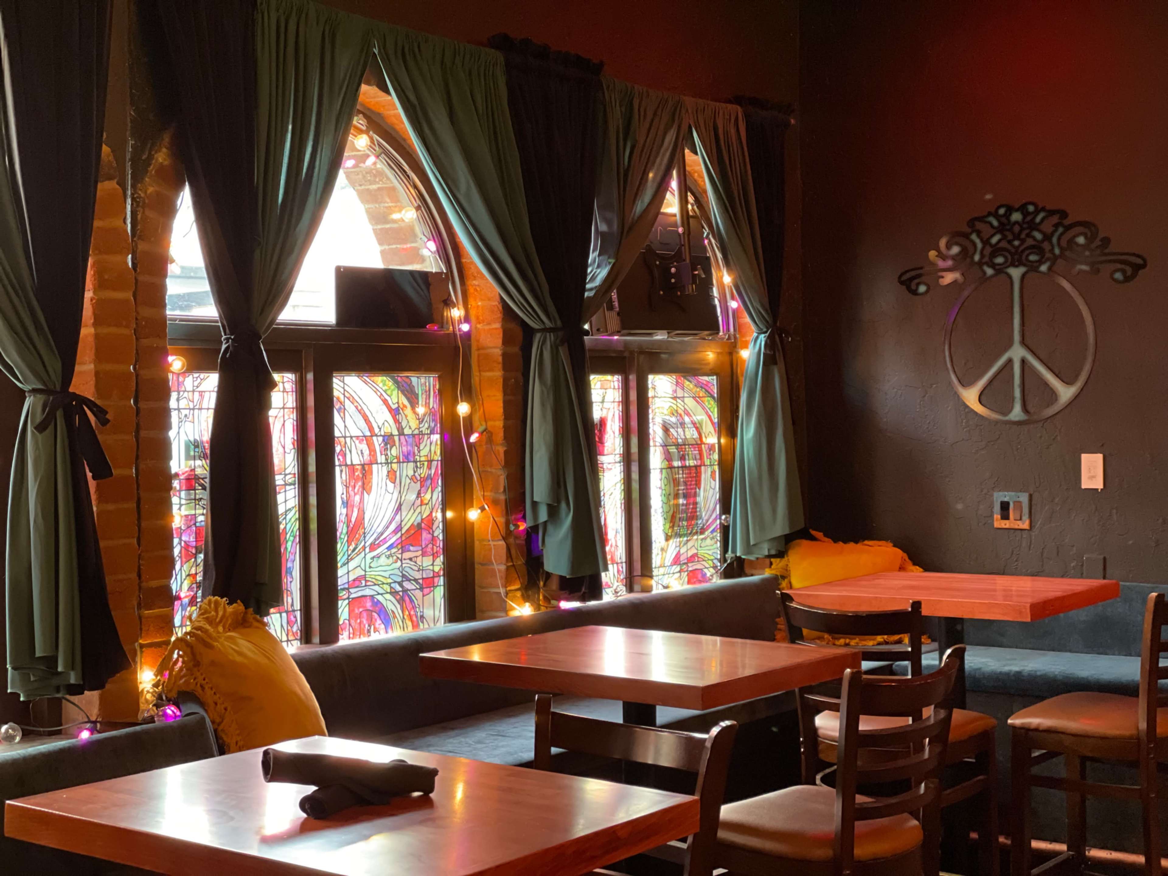 The interior of a dimly lit café features stained glass windows, tables, and chairs arranged near the windows adorned with curtains and decorative lighting.