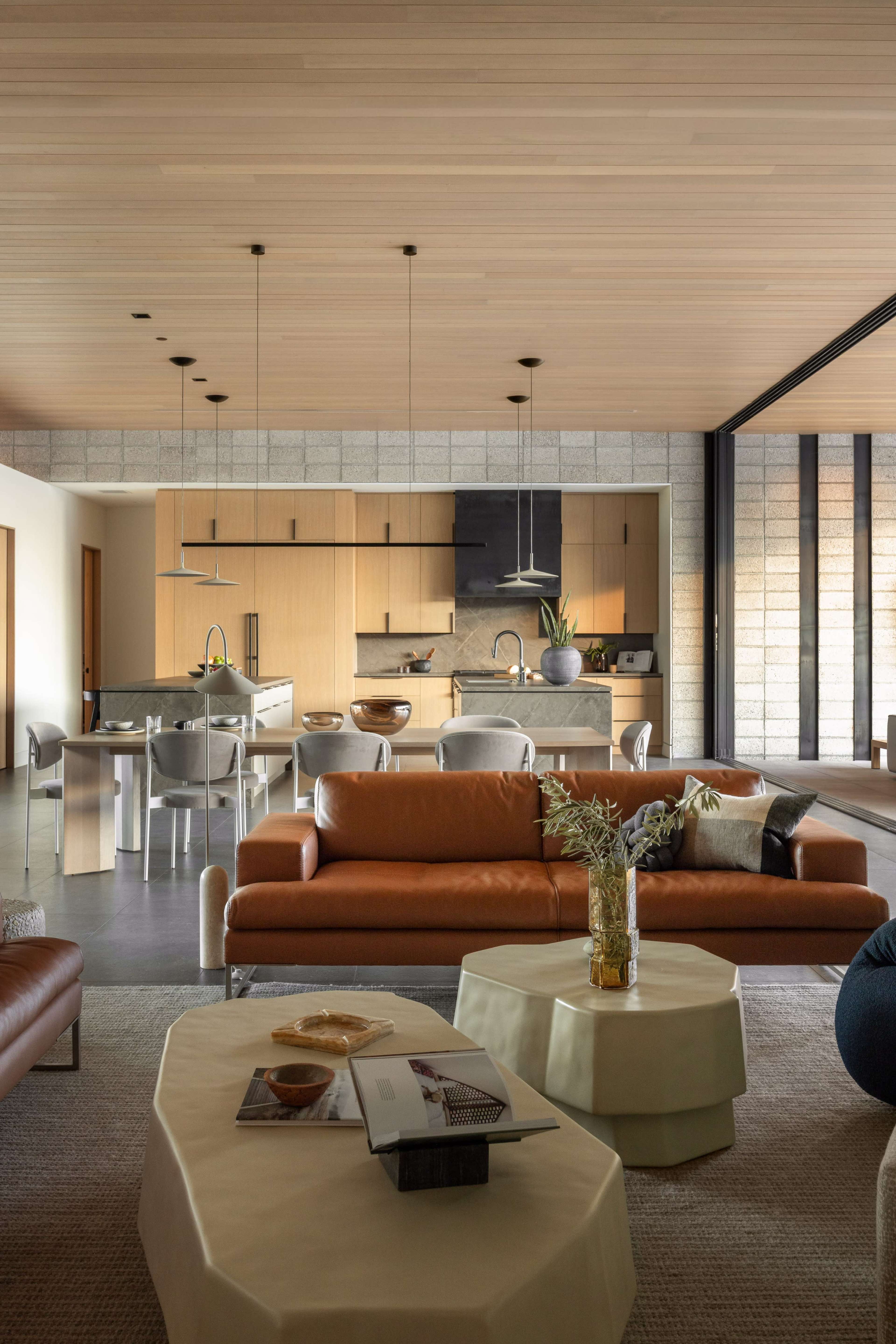 The image shows a modern living space with a leather sofa, a round coffee table, and an open kitchen area featuring wood cabinetry and metal appliances.