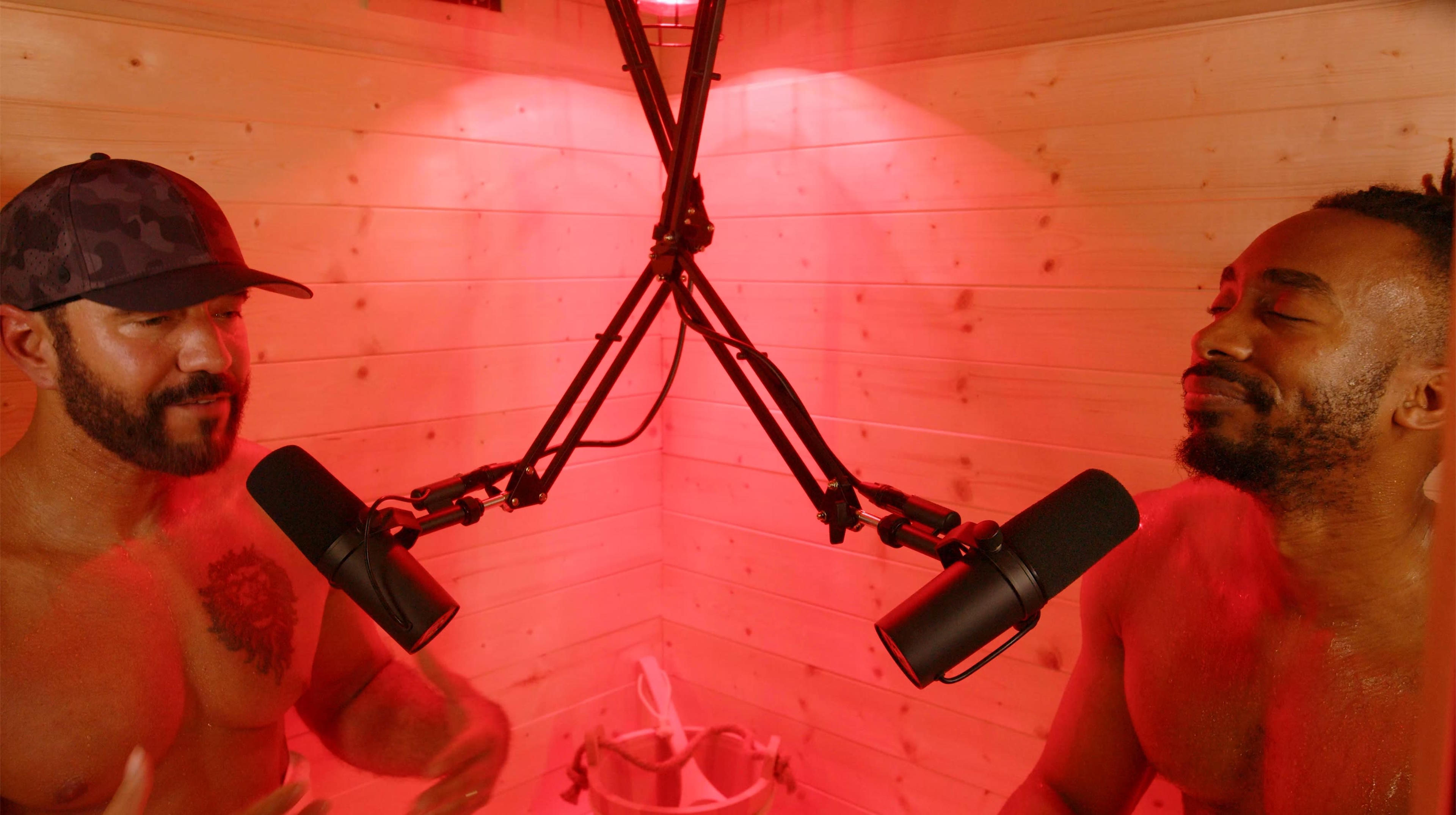 Two shirtless men are sitting in a red-lit sauna, each speaking into a microphone mounted on adjustable arms.