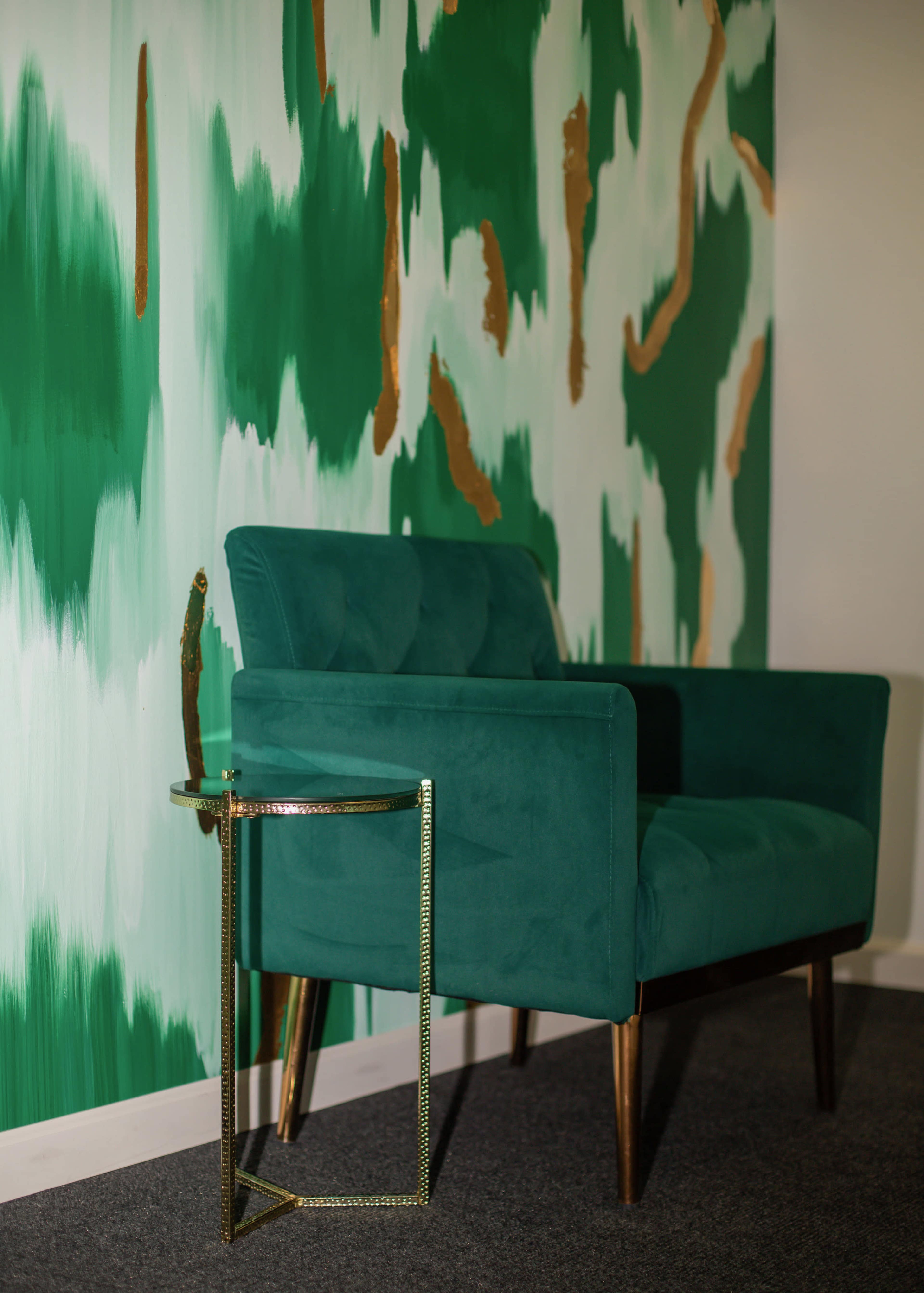 A teal upholstered chair is positioned next to a small gold side table, against a green and gold patterned wall.