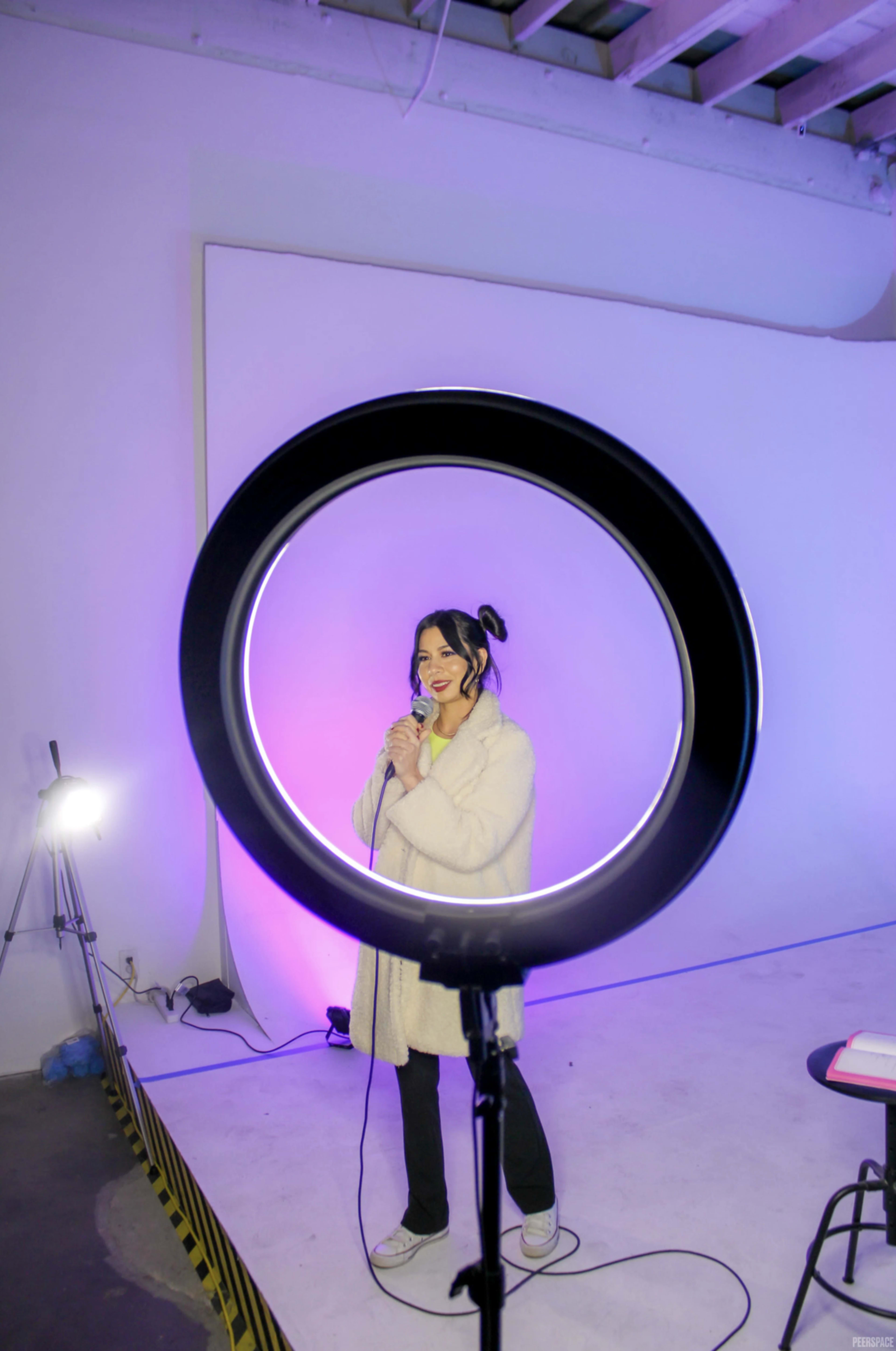 A woman stands in front of a microphone, illuminated by a ring light, in a studio with a white backdrop.