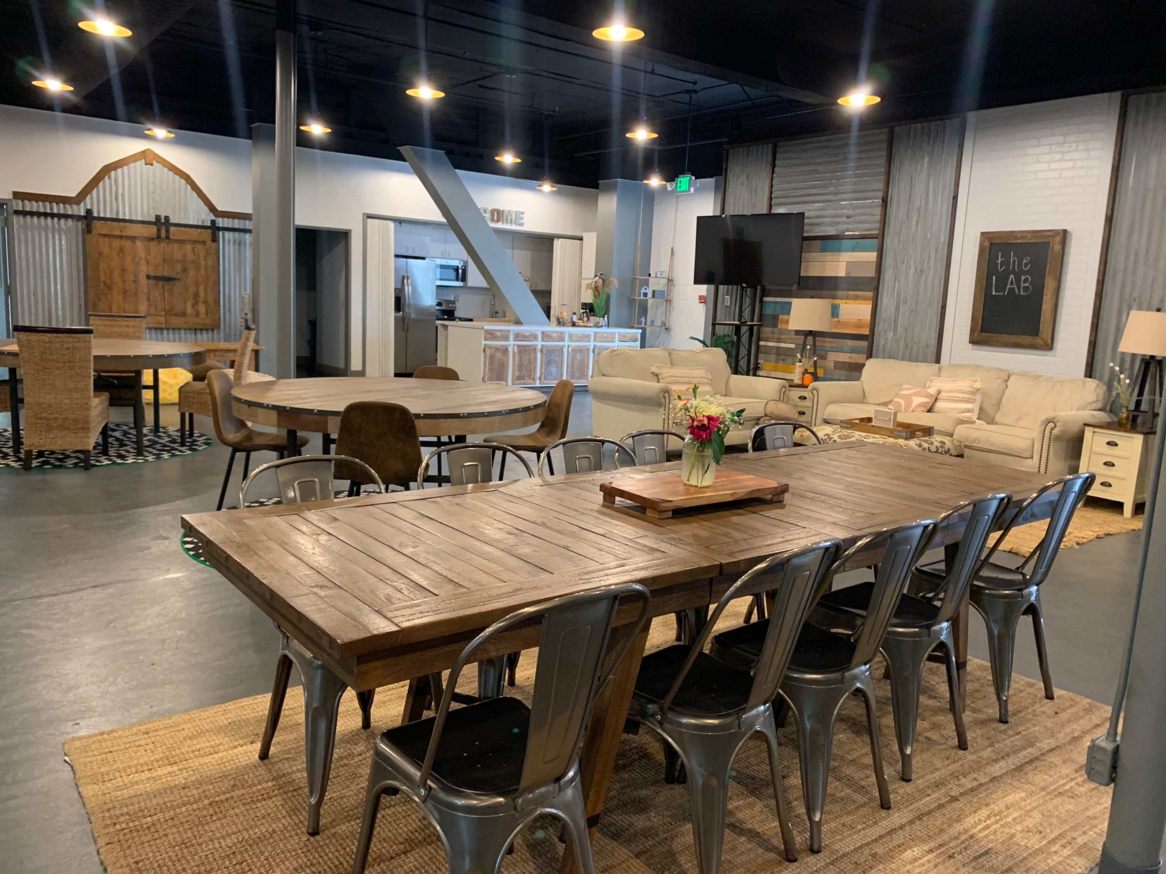 The image shows a modern, open space with a large wooden dining table, seating arrangements, a kitchen area visible in the background, and lounge furniture on one side.