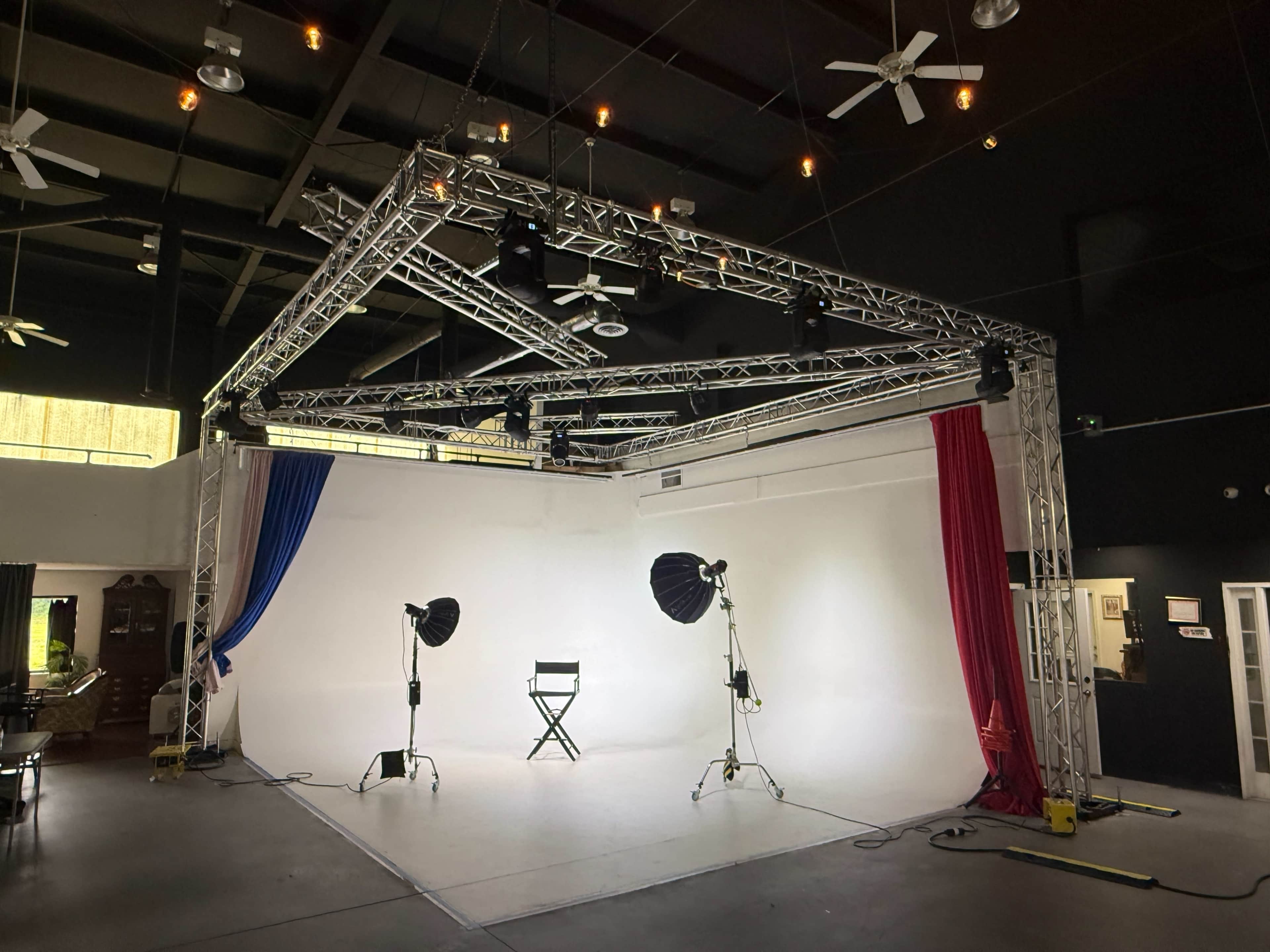 The image shows a spacious photography studio with a white backdrop, two studio lights set up on stands, and a black director's chair in the center.